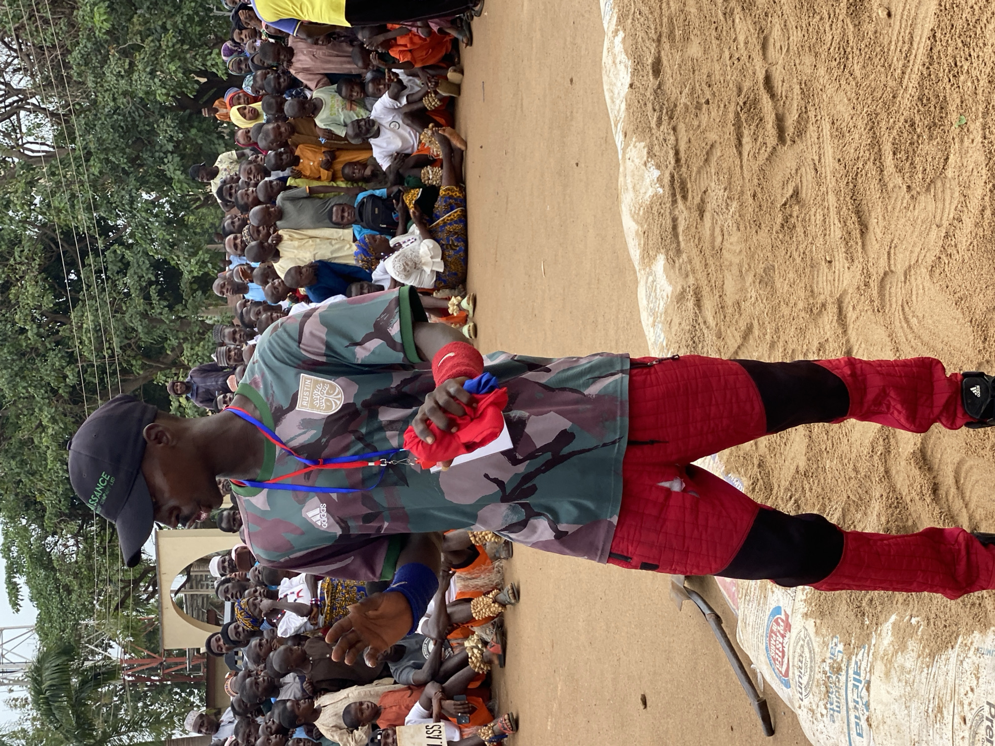 The image shows a man standing in a sandy area, possibly a wrestling arena. He wears a camouflage shirt with "AUSTIN" visible on the left breast and the Adidas logo on the right. He also wears red quilted pants with black accents and a black baseball cap with the word "RENAISSANCE" on it. He has a lanyard around his neck with blue and red stripes and is holding a red cloth in his left hand. His right arm has a blue wristband.
Behind the man, a large crowd of people is gathered. Some are seated, while others are standing. The crowd includes both men and women, and they are wearing various types of clothing. A white building is visible in the background behind the crowd.
To the right of the man is a large pile of sand and a partially visible white bag. There is a shovel leaning against the bag.
The image appears to have been taken in Kafanchan A, Nigeria, based on the clothing worn by people in the crowd. The scene suggests a cultural event, likely a wrestling match.