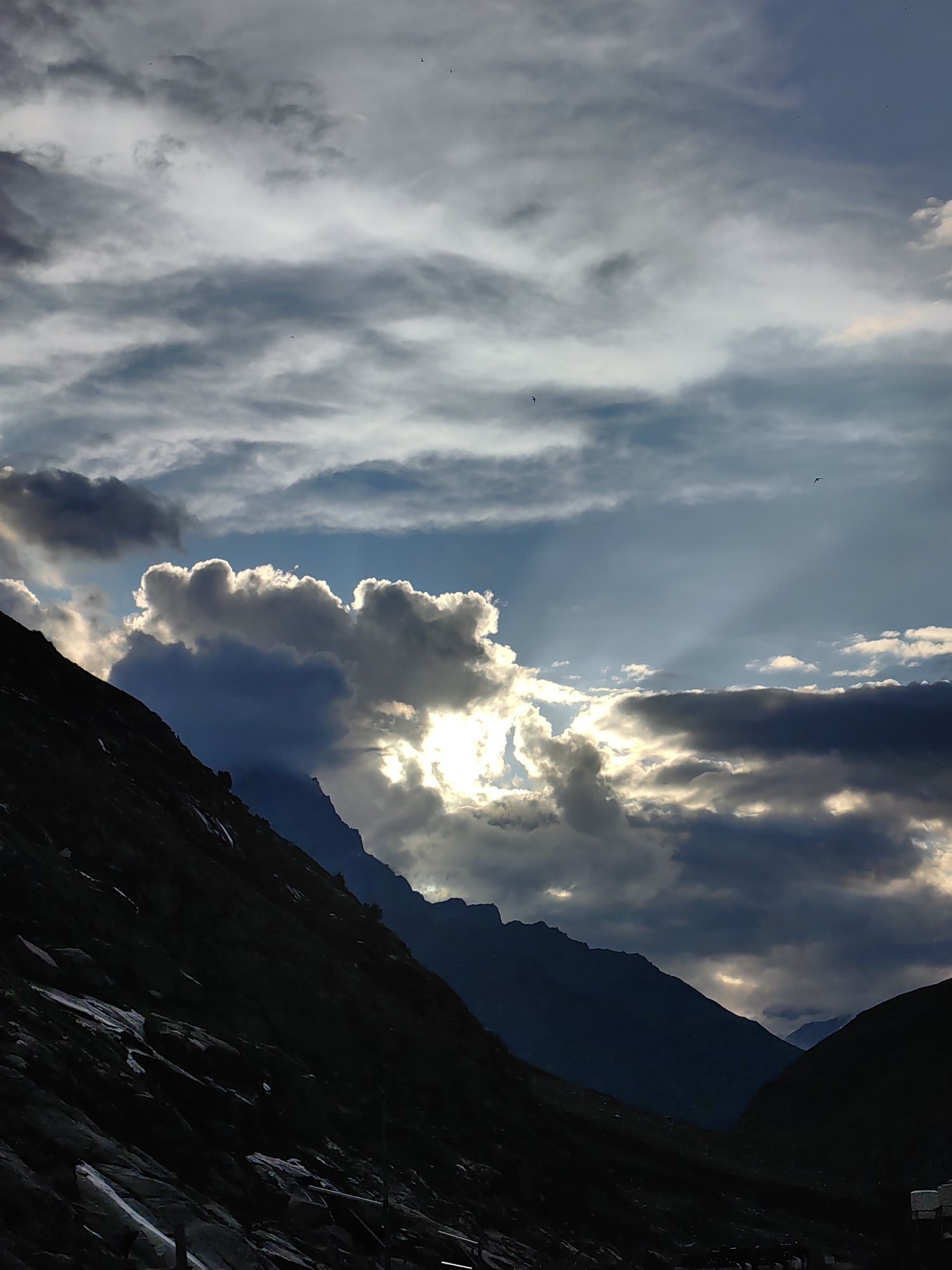 The image depicts a dramatic, cloudy sky over a mountainous landscape in Damphug, India. Sunlight pierces through gaps in the thick, grey clouds, casting bright beams onto the scene. The foreground is dominated by the dark silhouette of a steep, rocky mountain slope. In the background, several other mountain peaks are visible, fading into the distance. A few small birds are present in the sky. The overall mood is one of natural grandeur and a dynamic interplay of light and shadow.