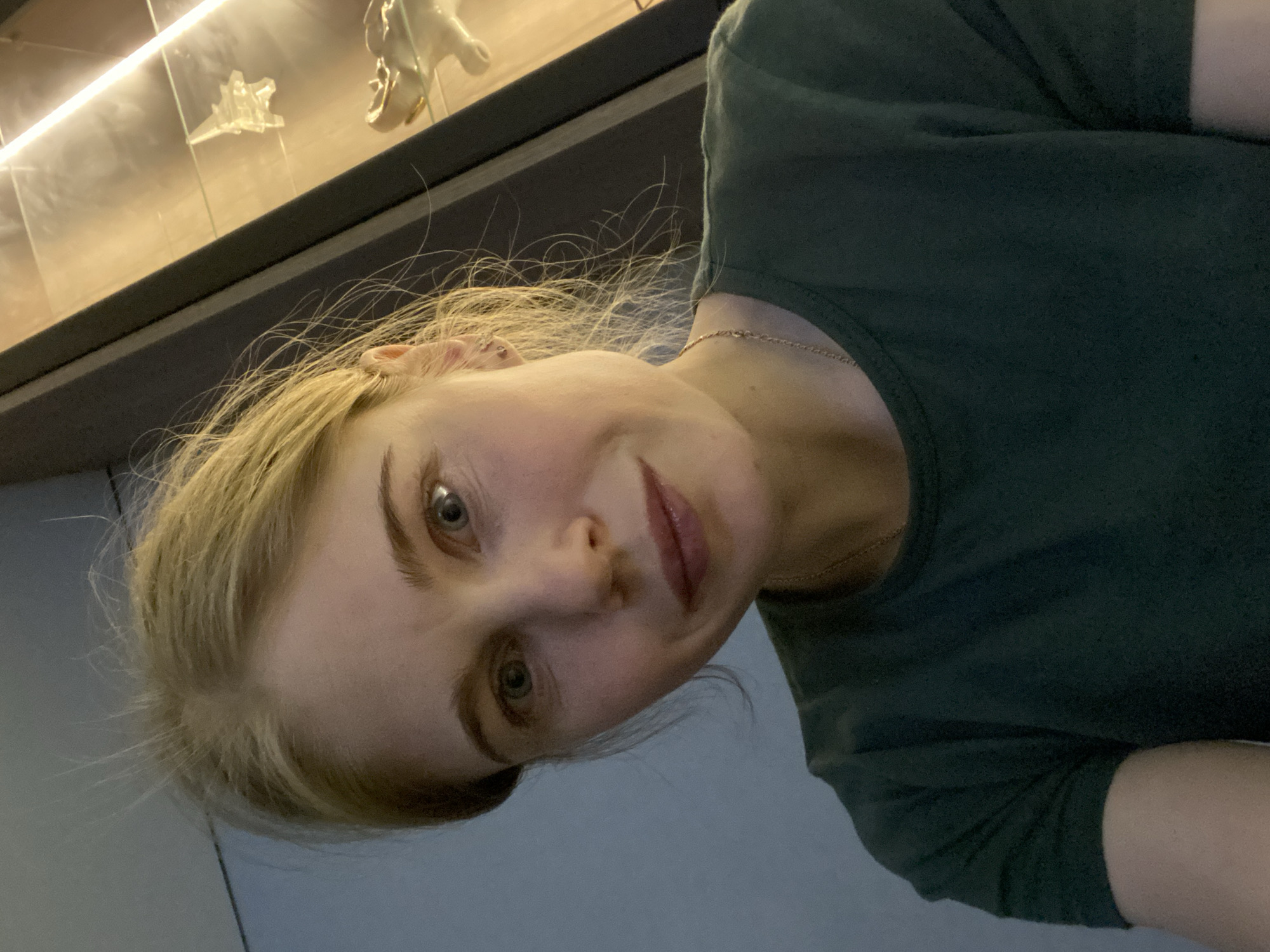 A woman with blonde hair and blue eyes is looking at the camera. She is wearing a dark green shirt. Behind her is a glass display case with illuminated objects, possibly miniatures. The location is potentially within the city of Chaikovsky, Russia, though there are no explicit indicators in the image.
