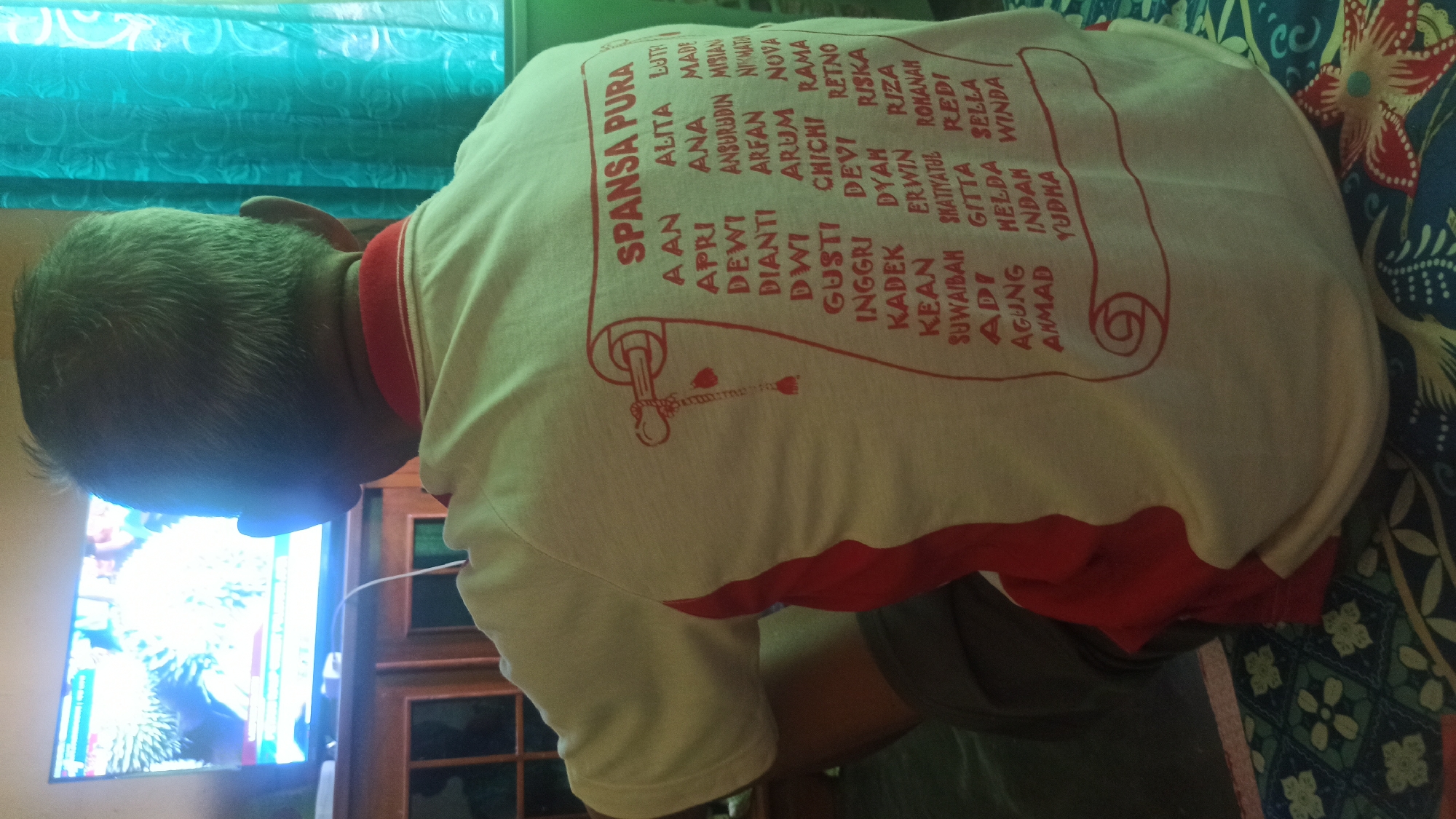 A medium shot captures an elderly Indonesian man from the back, seated in what appears to be a living room. He is facing a television screen displaying a nature documentary, its bright light illuminating his grey hair and the back of his head. He wears a white t-shirt with a red collar and red accents on the sides. The t-shirt has a red graphic print resembling a scroll with text in Indonesian, listing names. The names include "SPANSA PURA", "A AN", "APRI", "DEWI", "DIANTI", "GUSTI", "INGGRI", "KADEK", "KEAN", "SUWAIBAH", "ADI", "AGUNG", "ANMAD", "ALITA", "ANA", "ANSURUDDIN", "ARFAN", "ARUM", "CHICHI", "DEVI", "DYAH", "ERWIN", "SHATIYATUL", "GITT", "HELDA", "INDAH", "YUDHA", "LUTH", "MADE", "MISIAN", "NIKMATA", "NOVA", "RAMA", "RETNO", "RISKA", "RIZA", "ROHANA", "REDI", "SELLA", and "WINDA".
The man is seated on a bed or sofa covered with a patterned blue and green fabric. To the right of the man, a teal-colored curtain with an ornate pattern is visible. The environment suggests a domestic setting, likely a home in Tuba, Indonesia, given the Indonesian language on the t-shirt and the general ambiance. The man's posture indicates he is engrossed in watching the television.