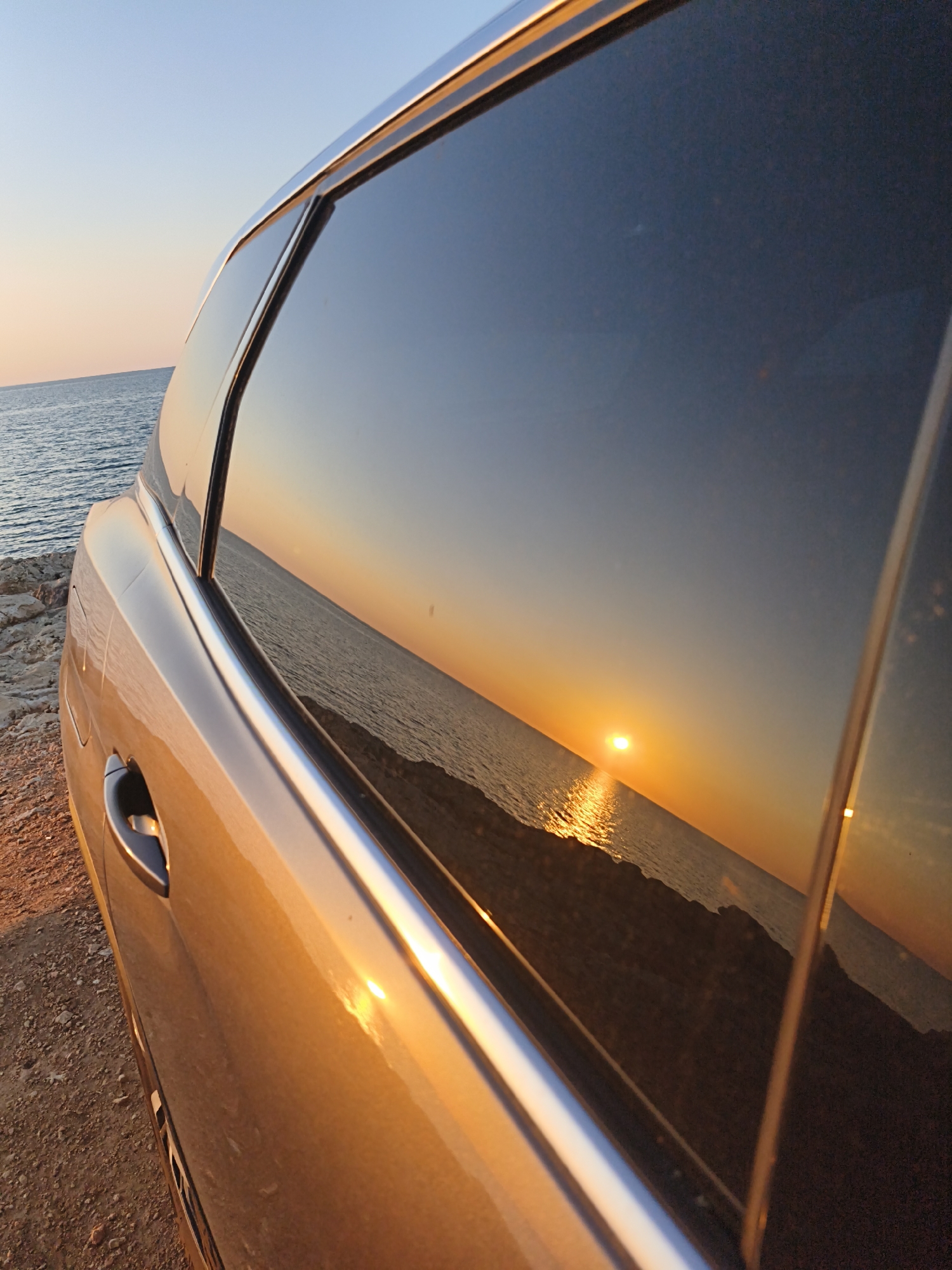 The image captures a golden-brown car parked on a rocky shore, offering a unique perspective of a sunset over the sea near Sevastopol, Ukraine. The primary focus is the vivid reflection of the setting sun and the horizon on the dark, tinted rear passenger window of the vehicle.
Within the reflection, a brilliant orange sun, low on the horizon, casts a radiant, shimmering path of light across the calm sea. The reflected sky above the sun transitions from fiery orange and yellow hues to a softer, clear blue at the top. On the actual scene outside the reflection, the calm sea is visible to the left, under a pale blue sky that subtly blends into a light orange closer to the horizon.
The car's metallic body, specifically the side panel and door handle, glows with the warm, golden light of the sunset, contrasting with the dark window. Silver trim outlines the window frame, catching the light. The ground next to the car is unpaved and consists of rocks and dirt. The time of day is clearly sunset, indicated by the low sun and warm, atmospheric lighting. The scene evokes a sense of peace and natural beauty. No text is visible in the image.