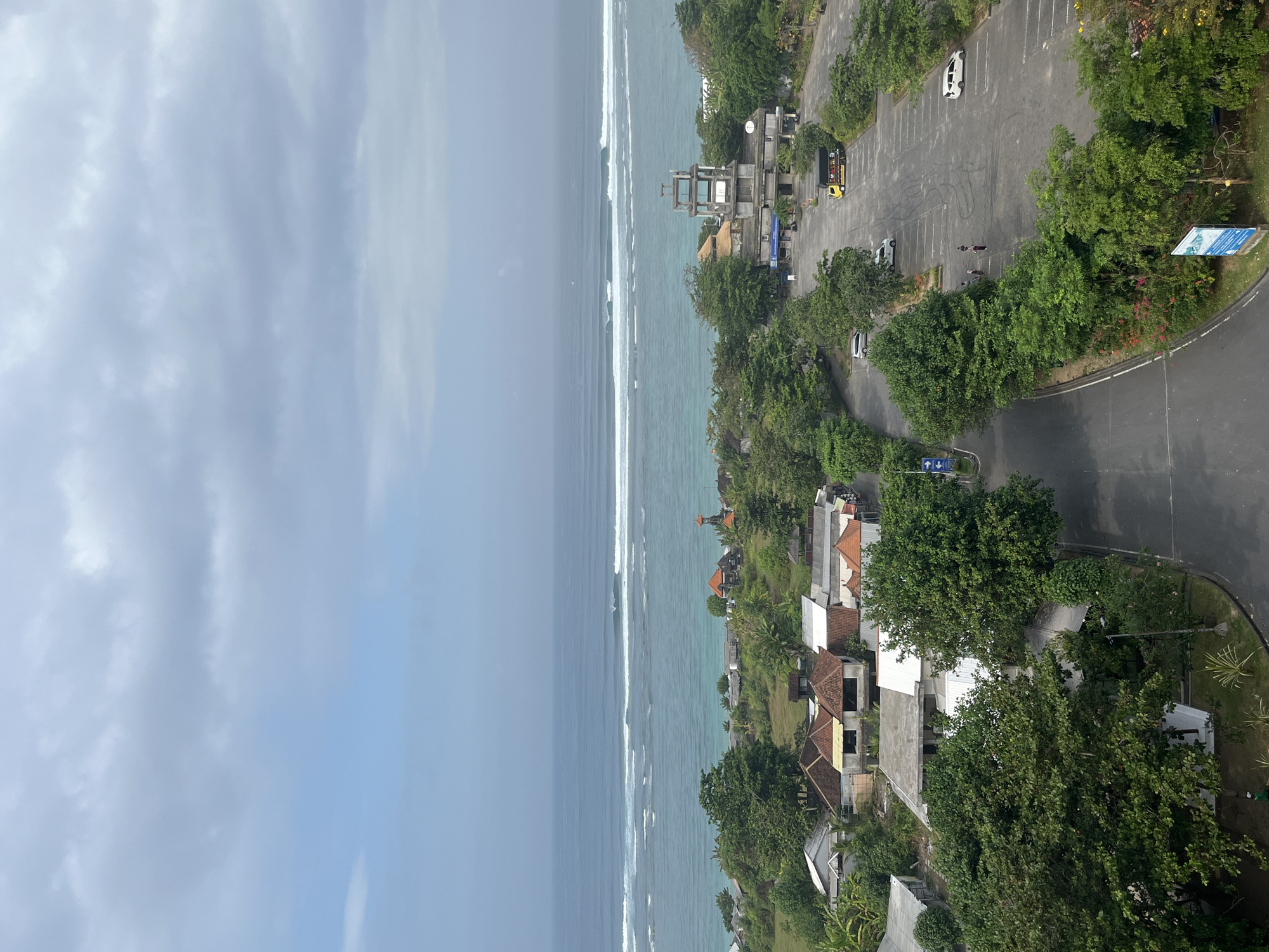 The image shows an elevated view of a coastal area in Kuta Selatan, Indonesia, with the ocean in the background and a developed area in the foreground. The ocean is a vibrant turquoise color with several sets of waves rolling towards the shore, indicating a popular surfing spot. Along the coastline, there are lush green trees and a few buildings with traditional Balinese architecture, featuring orange tiled roofs.
In the immediate foreground, a large paved parking lot is visible, with several vehicles parked, including a white car, a silver car, and a yellow truck with a black cargo bed. A road curves through the parking lot, and there are a few individuals standing in groups, appearing to be pedestrians. Adjacent to the parking lot, there is a modern, multi-story concrete structure with an open framework at the top, possibly a viewing tower or observation deck. More buildings with various architectural styles are scattered amongst dense green foliage, and a pathway or road winds through the scene. The sky is overcast with scattered clouds, creating a diffused light.