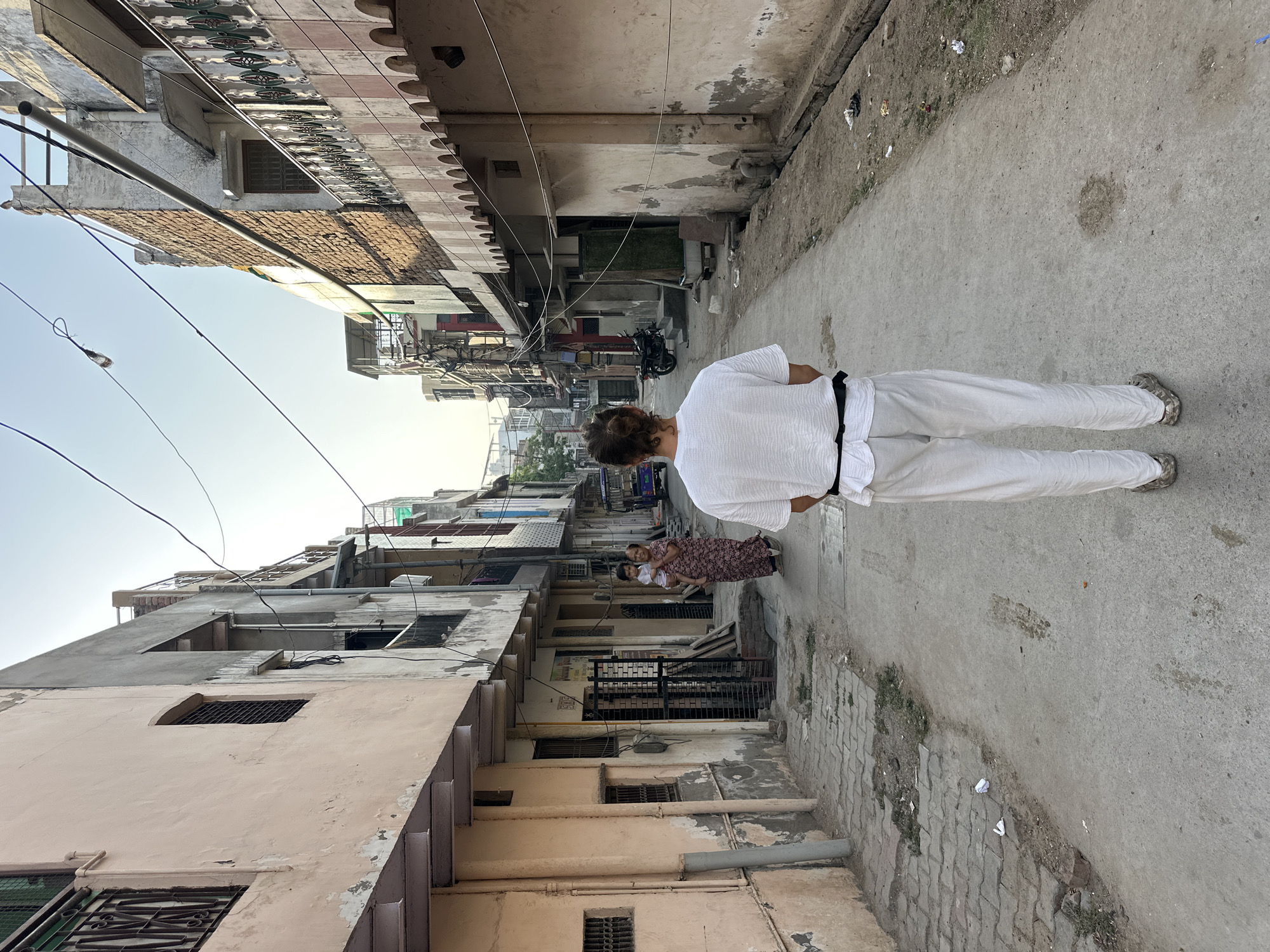 A street in Mathura, India, is depicted in a full shot. The street has light gray concrete on the right side and gray bricks on the left. Light gray concrete buildings line the street, with an older pink building on the left. Overhead, electrical wires traverse the street. A person dressed in a white outfit with a black belt walks away from the camera. In the background, an adult holds a child.