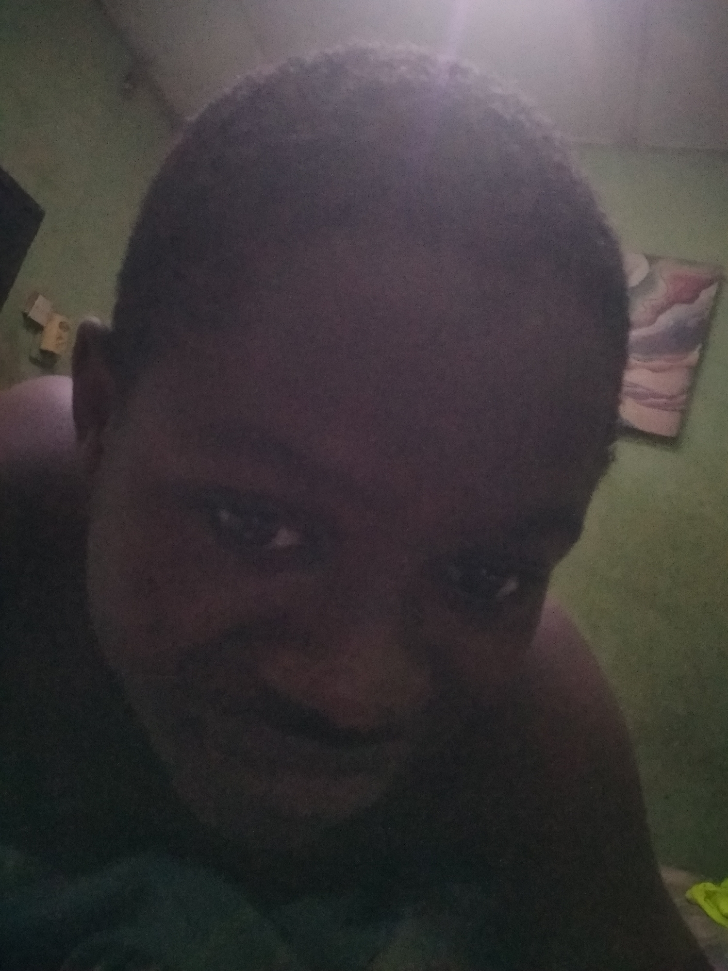 A close-up indoor selfie of a dark-skinned person with closely cropped hair, possibly male, is visible. The person is looking down towards the camera. The background includes a light green wall, a partially visible dark cabinet or shelf in the upper left corner, and a small, framed picture on the wall. Part of a green or yellow cloth is visible in the lower right corner of the frame. The image is dark and details are somewhat obscured. There is no verifiable information to confirm the location of the image is Shangisha, Nigeria.