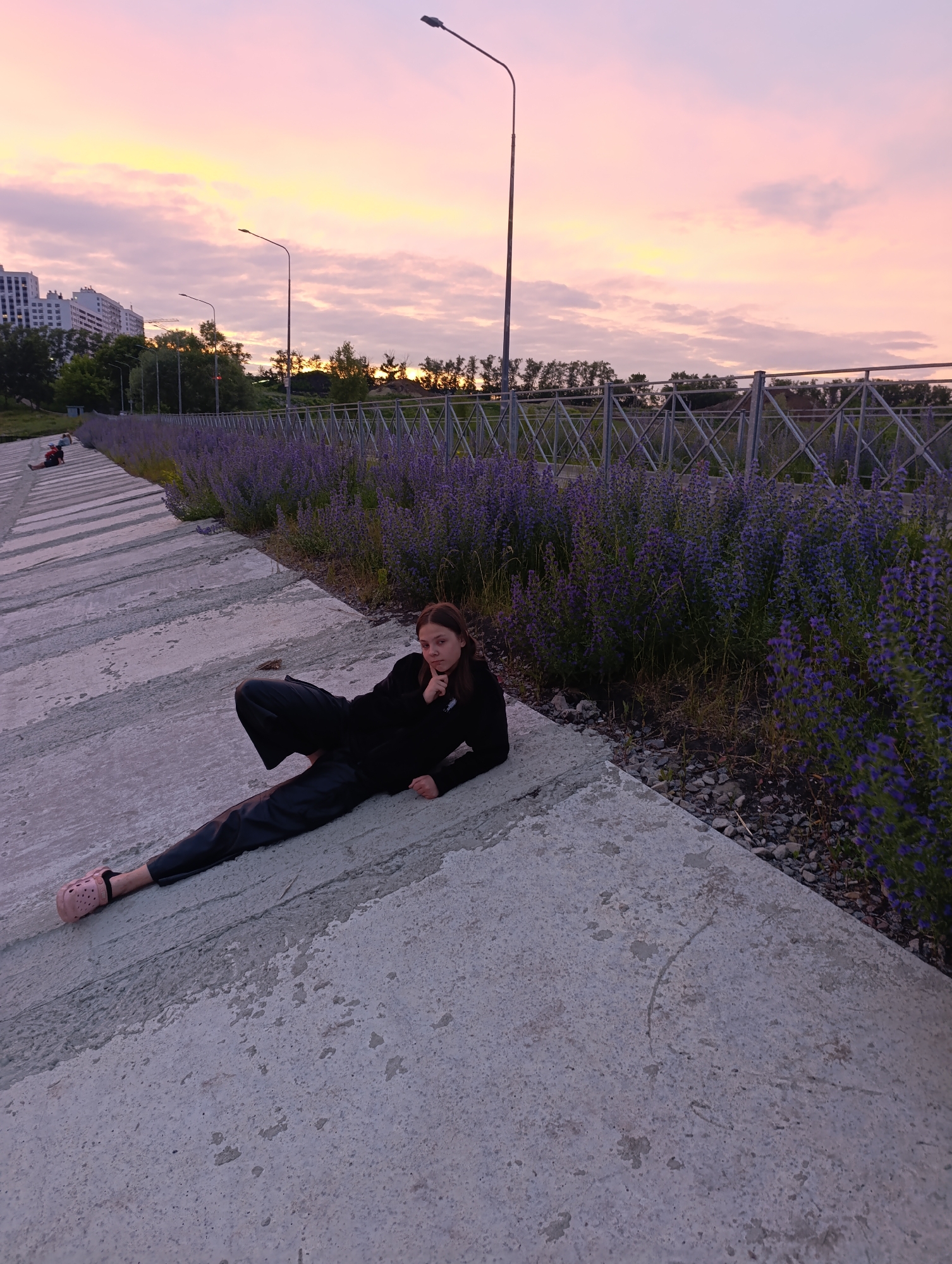 A young woman is lying on a concrete embankment overlooking a body of water, with a vibrant sunset painting the sky in shades of pink and orange. She is wearing a black top and black pants, with pink crocs on her feet. Her gaze is directed towards the camera, and her right hand is touching her chin. In the background, a row of purple flowers runs along a fence, and beyond that, trees and apartment buildings are visible. The concrete embankment is stepped, with other individuals seated further up the slope. The scene appears to be the embankment of the Sura River in Penza, Russia, a popular spot for recreation and enjoying the sunset.