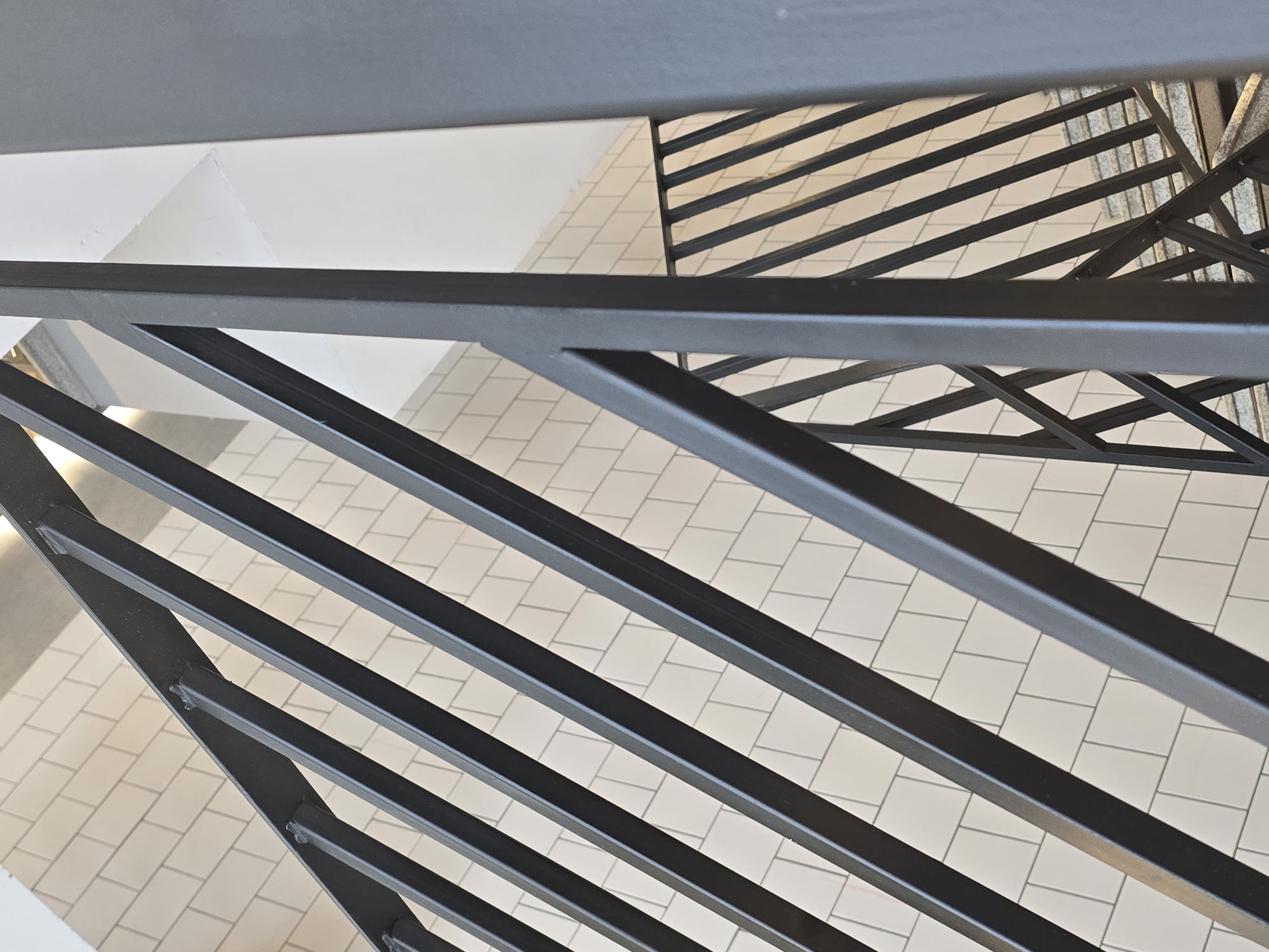 A high-angle shot depicts a metal railing with horizontal bars positioned over a tiled floor. The metal is dark, possibly black, and the tiles are light-colored, laid out in a grid pattern. A white wall is partially visible on the left side of the frame. The architectural setting appears to be indoors, possibly a public or commercial space.