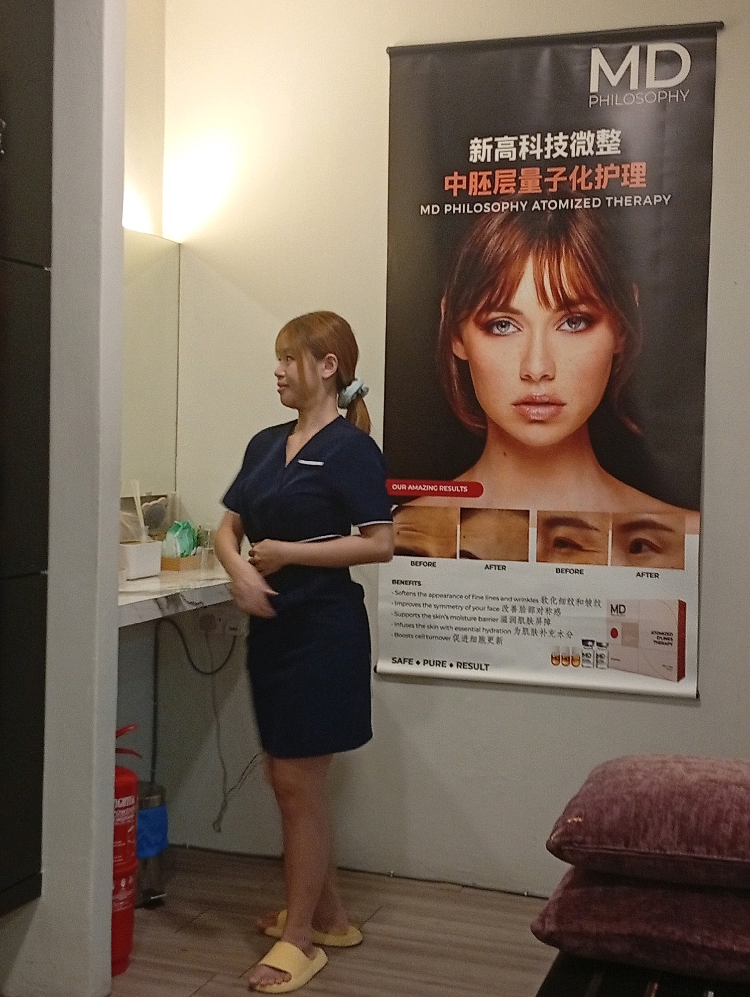 A woman with blonde hair tied in a ponytail is standing in a room, wearing a dark blue uniform. She is facing towards the left and looking slightly upwards. Behind her, a large banner with Chinese text and a woman's face is hanging on the wall. The banner advertises "MD PHILOSOPHY ATOMIZED THERAPY" and displays before-and-after images of facial treatments. To the left of the woman, there is a counter with some items on it, including what appears to be a white box and some green items. Further to the left, a red fire extinguisher is visible. The floor is tiled, and the walls are painted in a light color. The scene appears to be indoors, possibly in a salon or clinic. This location is in the city of Selayang Municipal Council, Malaysia.