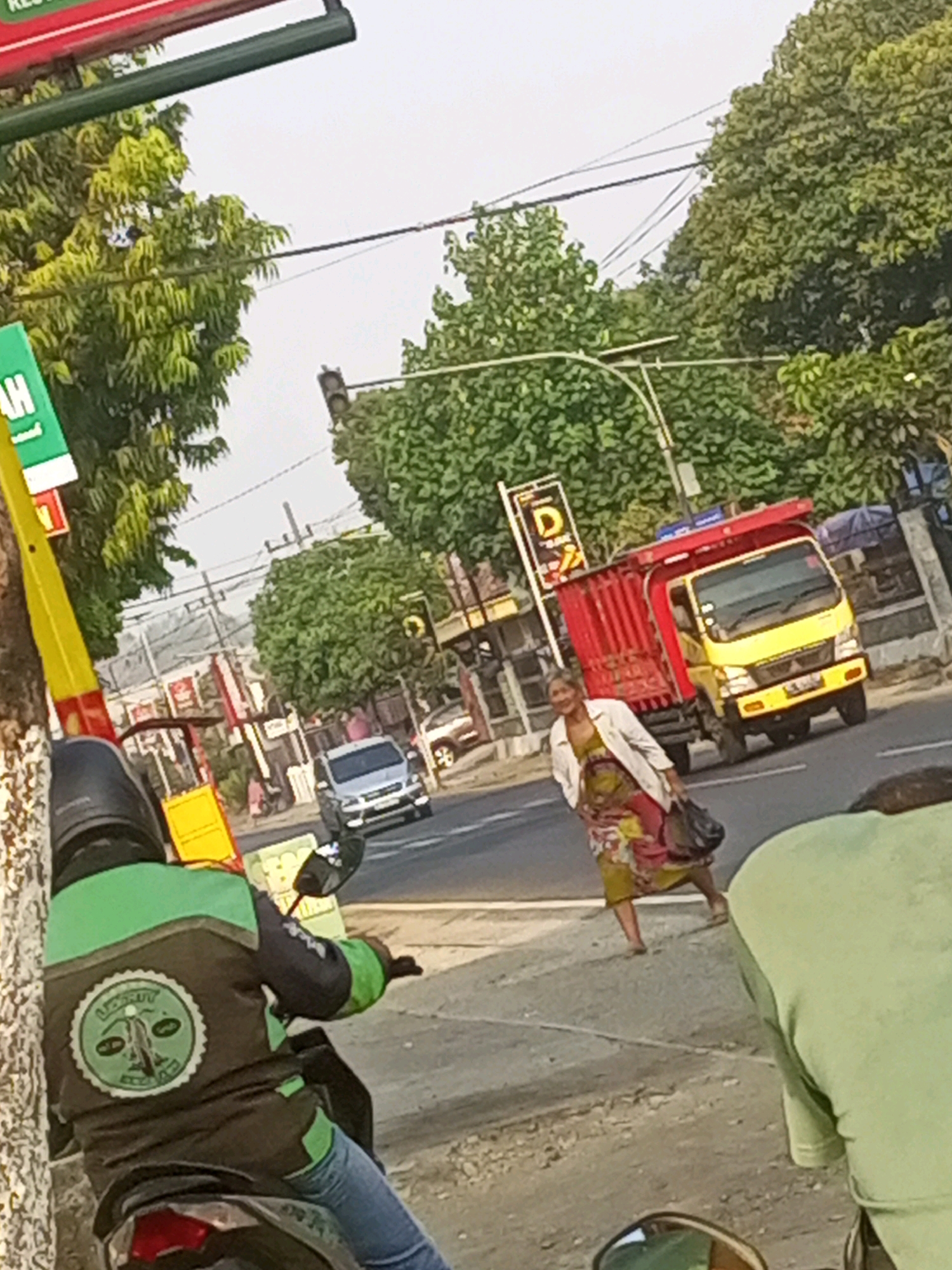 This is a photograph taken from a low angle, possibly from the perspective of someone riding a motorcycle in Gurah, Indonesia. In the foreground, the back of a person wearing a green and black jacket with a circular emblem is visible. They are wearing blue jeans and a helmet. The background shows a street scene with trees lining the side of the road. A yellow and red truck is stopped on the right side of the road. A woman in a colorful dress and white jacket is walking across the street in front of the truck, carrying a dark bag. A silver car is seen further down the road, and a traffic light is visible overhead. The sky is overcast.