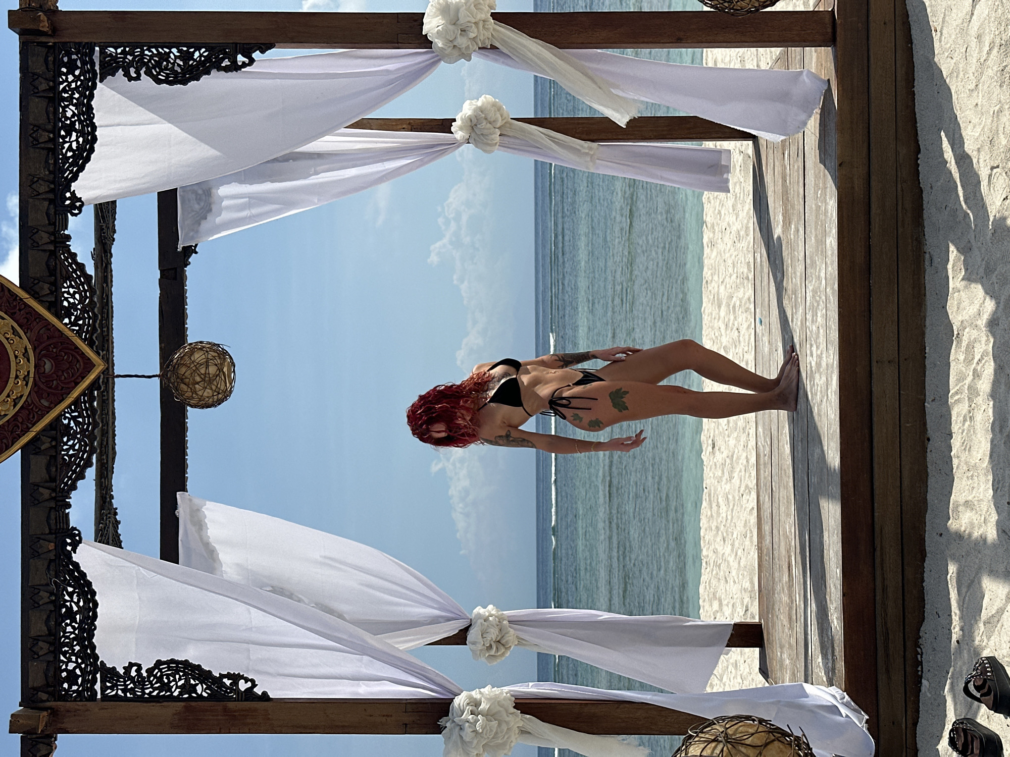 The image shows a woman on a wooden deck under a decorated structure, with an ocean backdrop. The woman, wearing a black bikini and sporting red hair, stands with her right arm extended and her left arm bent at the elbow. She has tattoos on her arms and legs.
The wooden deck is situated near a sandy beach, with the ocean stretching out to the horizon. The water appears calm with small waves.
The decorated structure overhead consists of wooden beams adorned with white fabric and floral embellishments. The structure is likely a gazebo or pavilion. Elaborate dark carved panels decorate parts of the structure.
The presence of the ocean and beach suggests a coastal location in Unknown, Indonesia. The time of day appears to be daytime, under clear or partly cloudy skies. A pair of shoes lie on the sand at the edge of the deck.