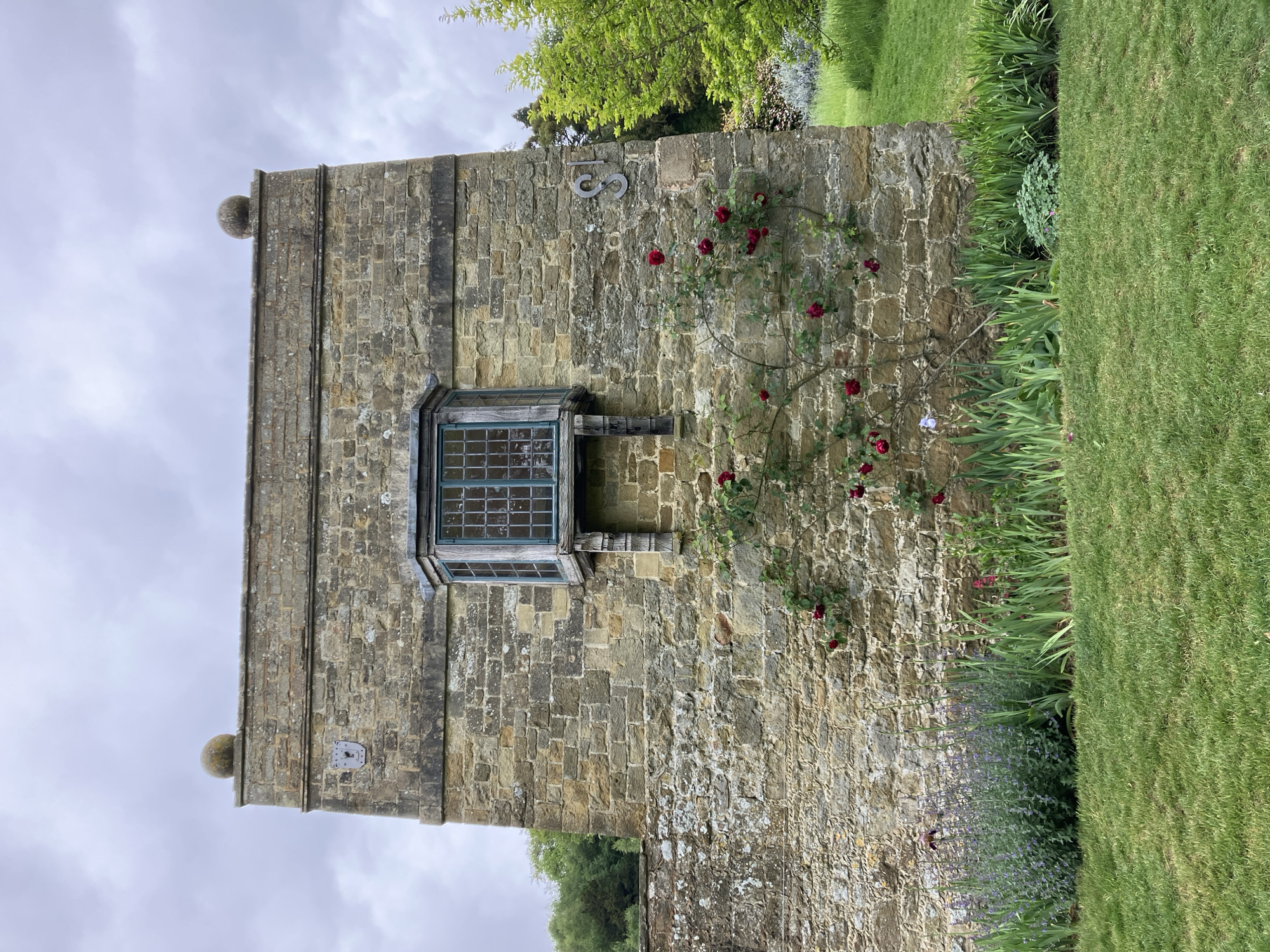 The image depicts a weathered stone wall, likely part of an old building or structure. The wall is constructed from irregularly shaped stones, with visible mortar lines. The image is taken in Sevenoaks, United Kingdom. A square window is set into the wall, with a greenish frame and multiple panes. A climbing rose bush with red flowers is growing on the right side of the wall, partially obscuring the stone surface. To the right of the wall, there is a strip of green grass, suggesting a garden or park setting. Above the window, there are metal numbers, the number 18. The top of the wall features a decorative ledge and two spherical objects at the corners. The sky is cloudy, with a soft light filtering through. The building is perpendicular to the horizon.