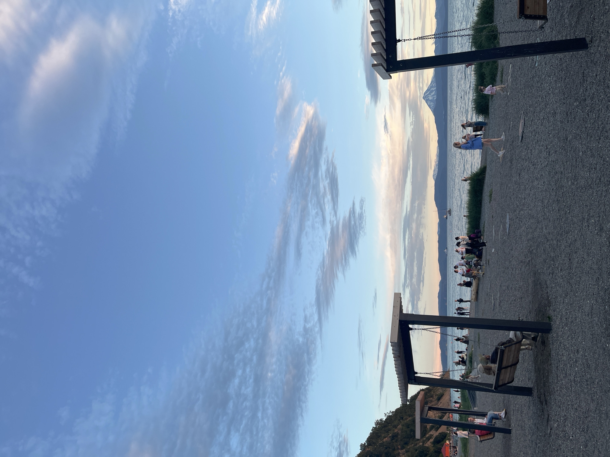 The image depicts a serene sunset scene on a gravelly beach in Petropavlovsk-Kamchatsky, Russia. The sky is a soft blend of blue and pink hues, with wispy clouds scattered across the expanse. In the foreground, two large wooden swings with comfortable seating are present, one occupied by a person. Further back, a group of people are gathered by the water, some sitting on the shore, others standing and conversing. The calm, vast body of water stretches towards the horizon, where a majestic snow-capped volcano dominates the landscape, its peak silhouetted against the fading light. A few sailboats are visible on the water, adding to the tranquil ambiance. The environment appears to be a popular recreational area, with lush green grass bordering the beach. The overall atmosphere is peaceful and idyllic, showcasing the natural beauty of the region.