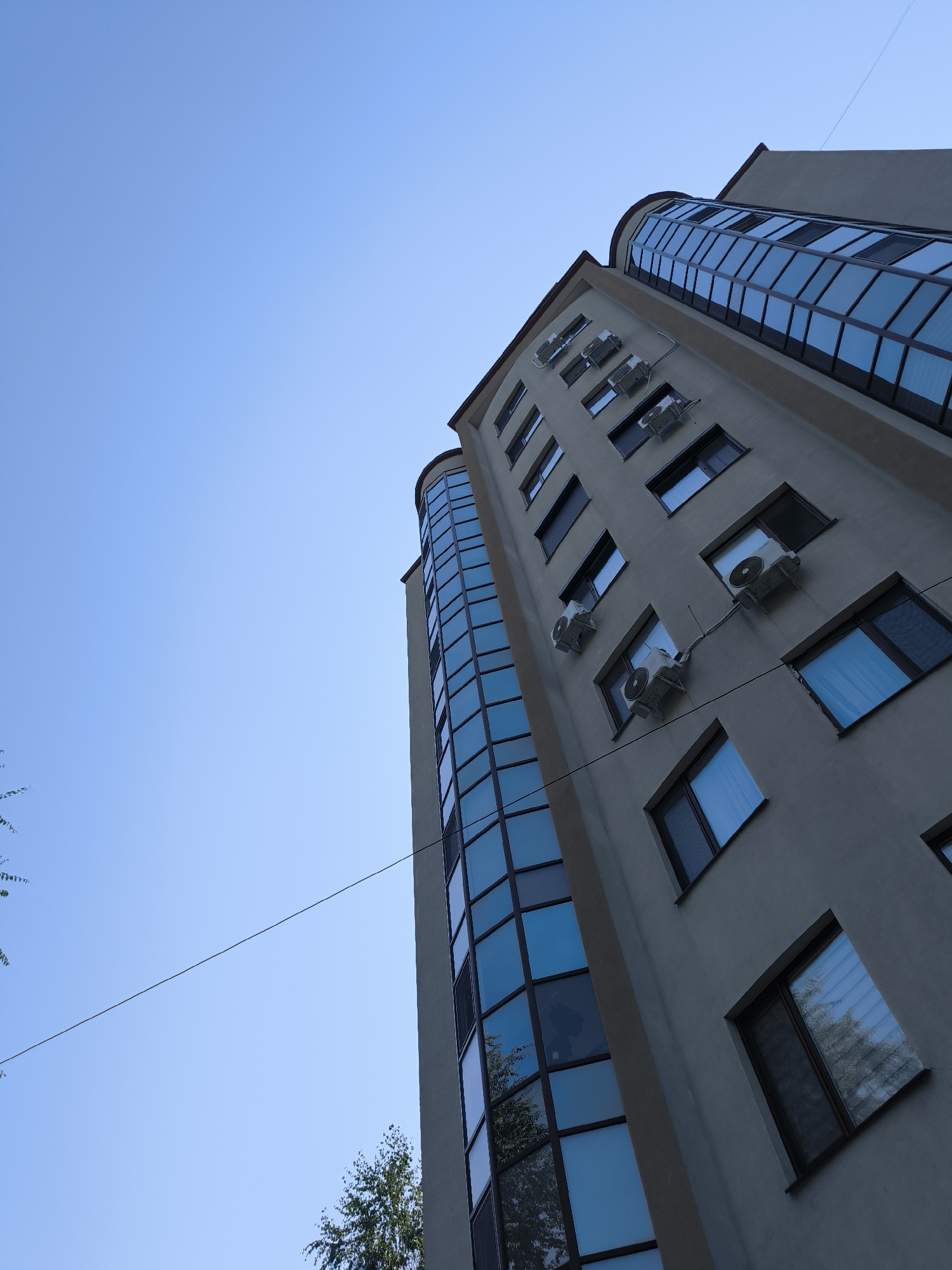 This image shows a low-angle view of a modern, multi-story building against a clear blue sky. The building has a light-colored facade and features large, reflective windows, some of which are curved, giving the building a contemporary architectural style. Multiple air conditioning units are visible mounted on the exterior walls. A thin wire stretches across the sky from the left side of the frame towards the building. Trees are visible in the lower left and lower right corners of the image, suggesting an urban or suburban setting. The perspective is looking upwards, emphasizing the height of the building and the vastness of the sky. This appears to be a residential or commercial building located in the city of Chișinău, Moldova.