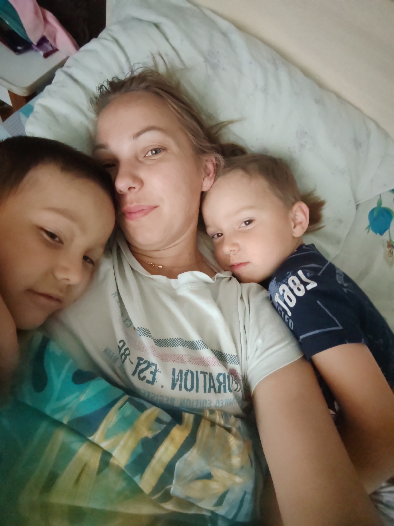 The image shows a close-up, overhead view of a woman and two young boys lying in bed. The woman is in the center, with one boy on her left and another on her right, both snuggled close to her. The woman has blonde hair and is looking at the camera with a slight smile. She is wearing a white t-shirt with text on it. The boy on the left has dark hair and is looking towards the camera. The boy on the right has lighter brown hair and is looking away from the camera, with his arm around the woman. They are all under a light-colored blanket with a subtle pattern. The background shows part of a bed with pillows and bedding. There is no discernible information in the image that would place the scene specifically in Чухлэм, Russia, or any other city.