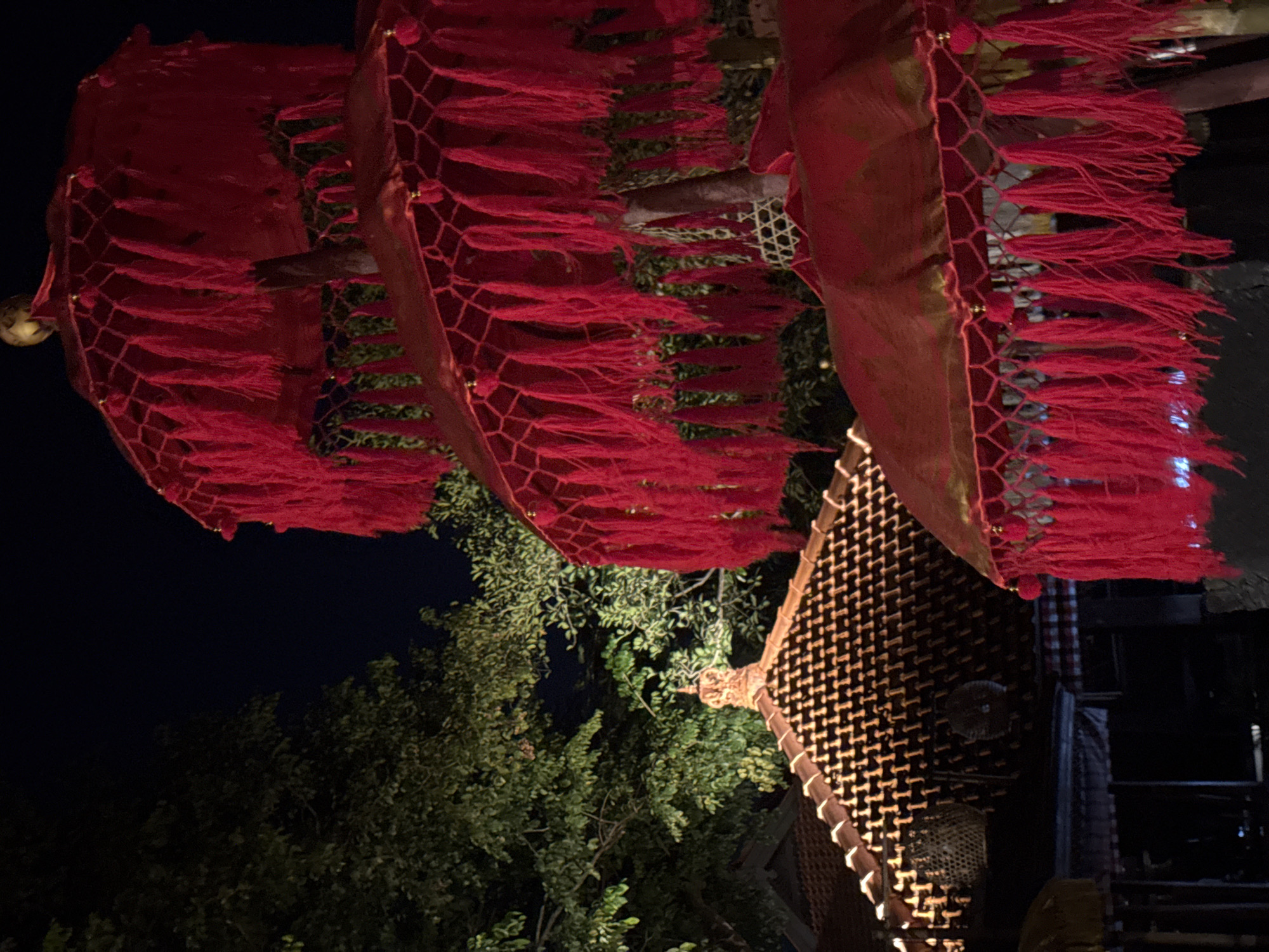 At night, the image shows an outdoor scene likely in Kuta Selatan, Indonesia, featuring decorative elements in the foreground. Above, a red hanging decoration with fringe is suspended, featuring a netted pattern at its edges. Below the hanging decoration are trees with green foliage. To the right is a structure with a woven or grid-like pattern, possibly part of a building or art installation. Beyond the structure, there is dark space suggesting shadows or further structures.