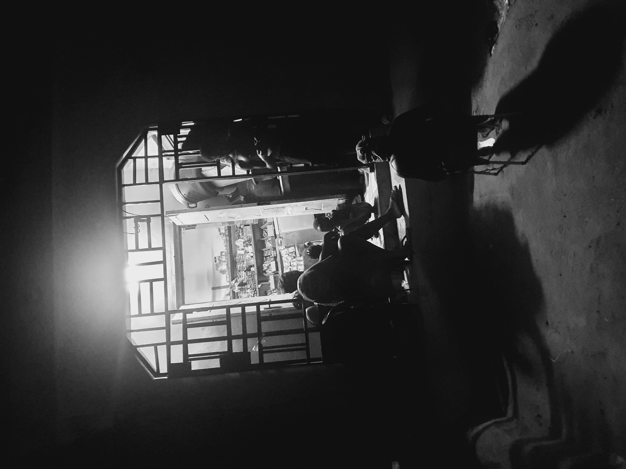 In the dark, grainy image, the outline of a shop is visible through a window with a metal frame. The shop displays shelves with merchandise. People appear to be inside the shop. Outside the shop, more individuals are sitting and standing. The location is suggested to be in Lokoja, Nigeria. The image captures a dimly lit, possibly nighttime, scene of a small commercial establishment with people inside and outside.