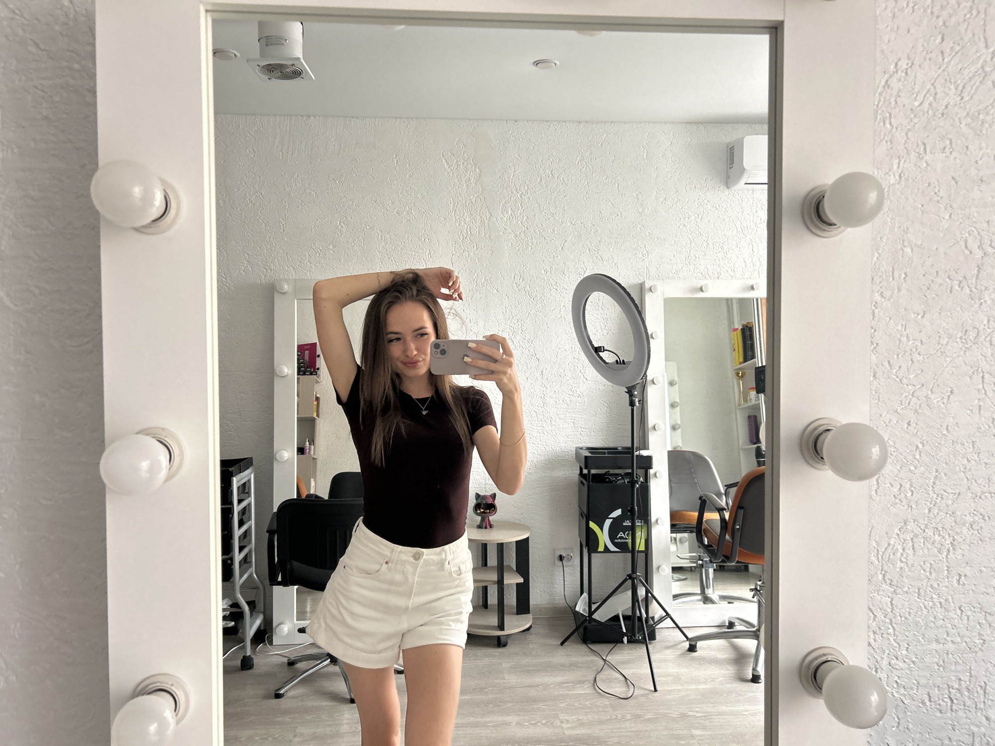 A young woman is taking a mirror selfie in a room. She is wearing a dark brown t-shirt and white shorts. Her right arm is raised, and she is holding her hair. She is holding a pink smartphone in her left hand. The mirror is framed with lights. In the background, there are salon chairs, a ring light on a tripod, and shelves with bottles and other items. The walls are textured and white.