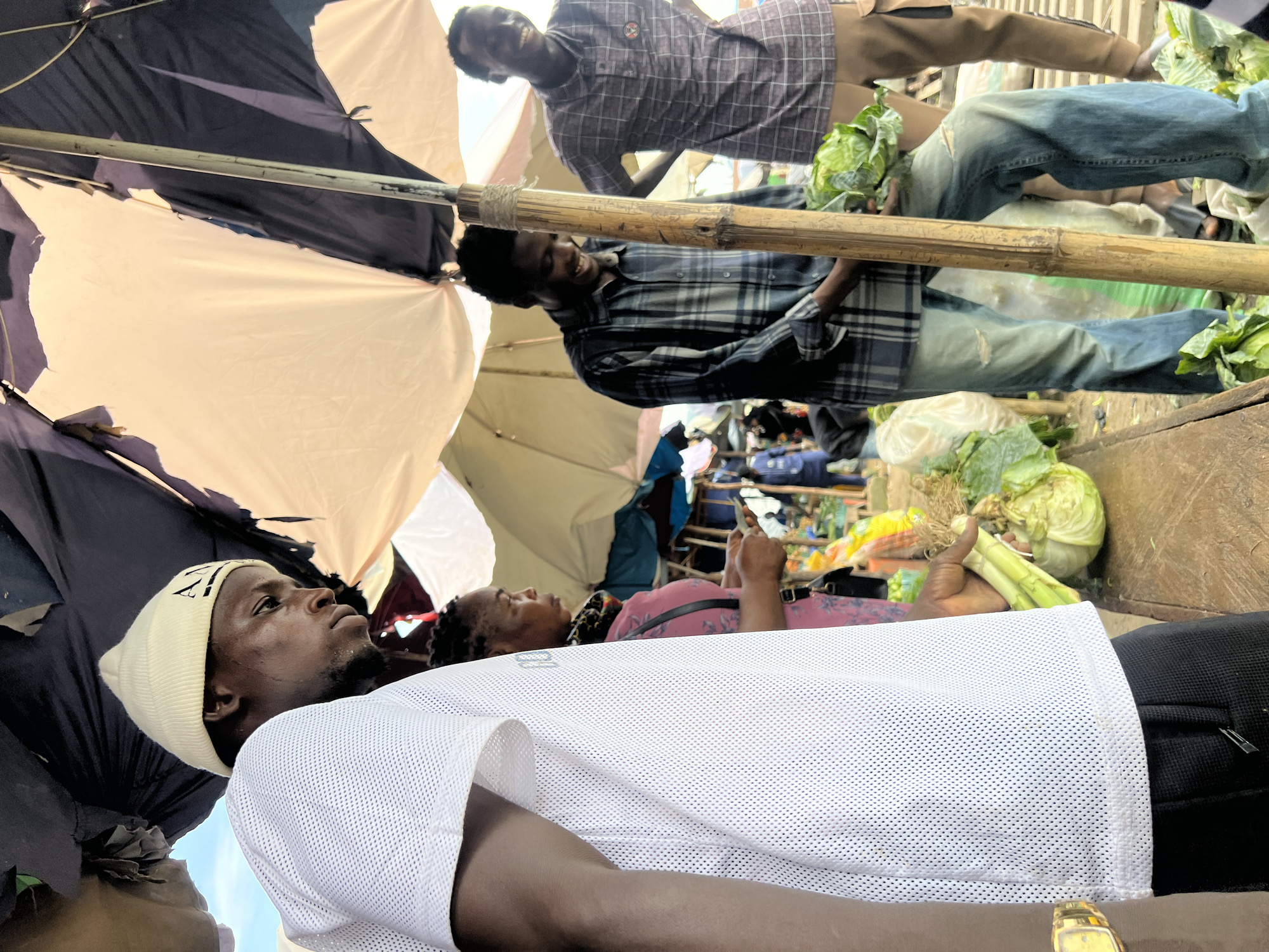 In Oko Amakom, Nigeria, a bustling market scene unfolds under a patched and torn umbrella. Four individuals are visible. In the foreground, a man wearing a white mesh t-shirt and black trousers, with a white beanie, is partially visible, turned to the right. To his left, a woman in a pink dress with a black and floral scarf around her neck, holding a phone, is looking down. Behind them, two men stand near a stall. The man on the left, wearing a blue and grey checkered shirt and ripped blue jeans, is smiling and holding a head of cabbage. To his right, another man in a purple and grey checkered shirt and tan trousers, is also smiling, his hands on his hips. The market environment is characterized by wooden stalls laden with fresh produce, including cabbages and other leafy greens. The overall impression is one of vibrant daily commerce in a Nigerian city.