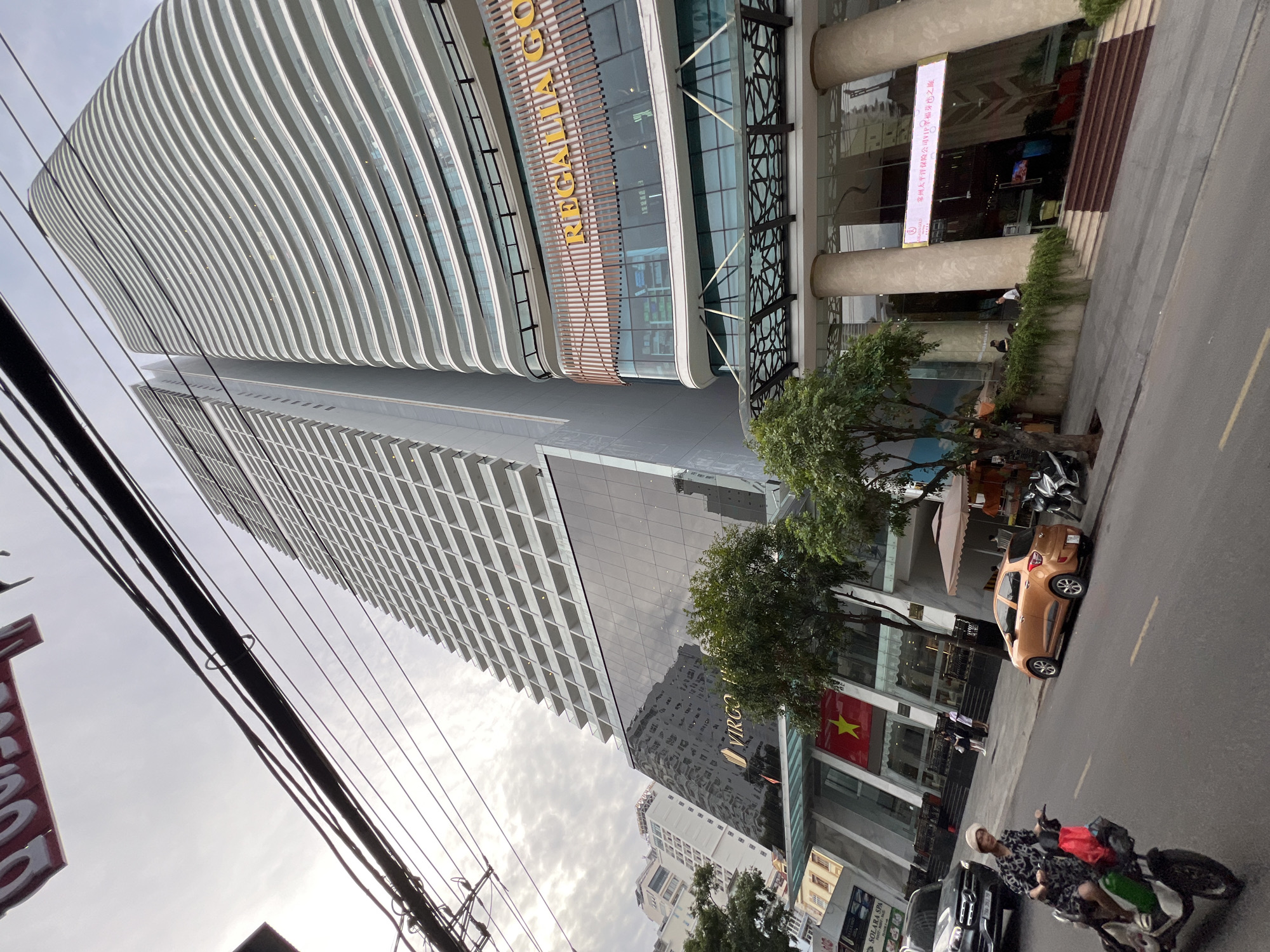 The image depicts a street scene in Nha Trang, Vietnam, dominated by two tall, modern buildings. The building on the right features a distinctive wavy facade with numerous balconies. Above its entrance, a sign reads "REGALIA GOLD" in large, golden letters. The building on the left is a more conventional skyscraper with a grid-like pattern of balconies and windows. A large glass facade on this building reflects the surrounding environment.
In the foreground, a woman wearing a hat and a patterned dress rides a white scooter down the street. She is carrying a red bag and a green bag on the scooter. To her right, an orange car is parked on the side of the road. Further down the street, several motorcycles are parked. A black Mercedes sedan is also visible on the left side of the street.
A Vietnamese flag hangs prominently on the left building, near the entrance. Lush green trees line the sidewalk in front of the buildings. The sky is overcast, with visible power lines stretching across the top of the frame. The street appears to be a commercial area, with shops and businesses visible at the base of the buildings. A digital billboard with Chinese characters is also visible above the entrance of the right building.