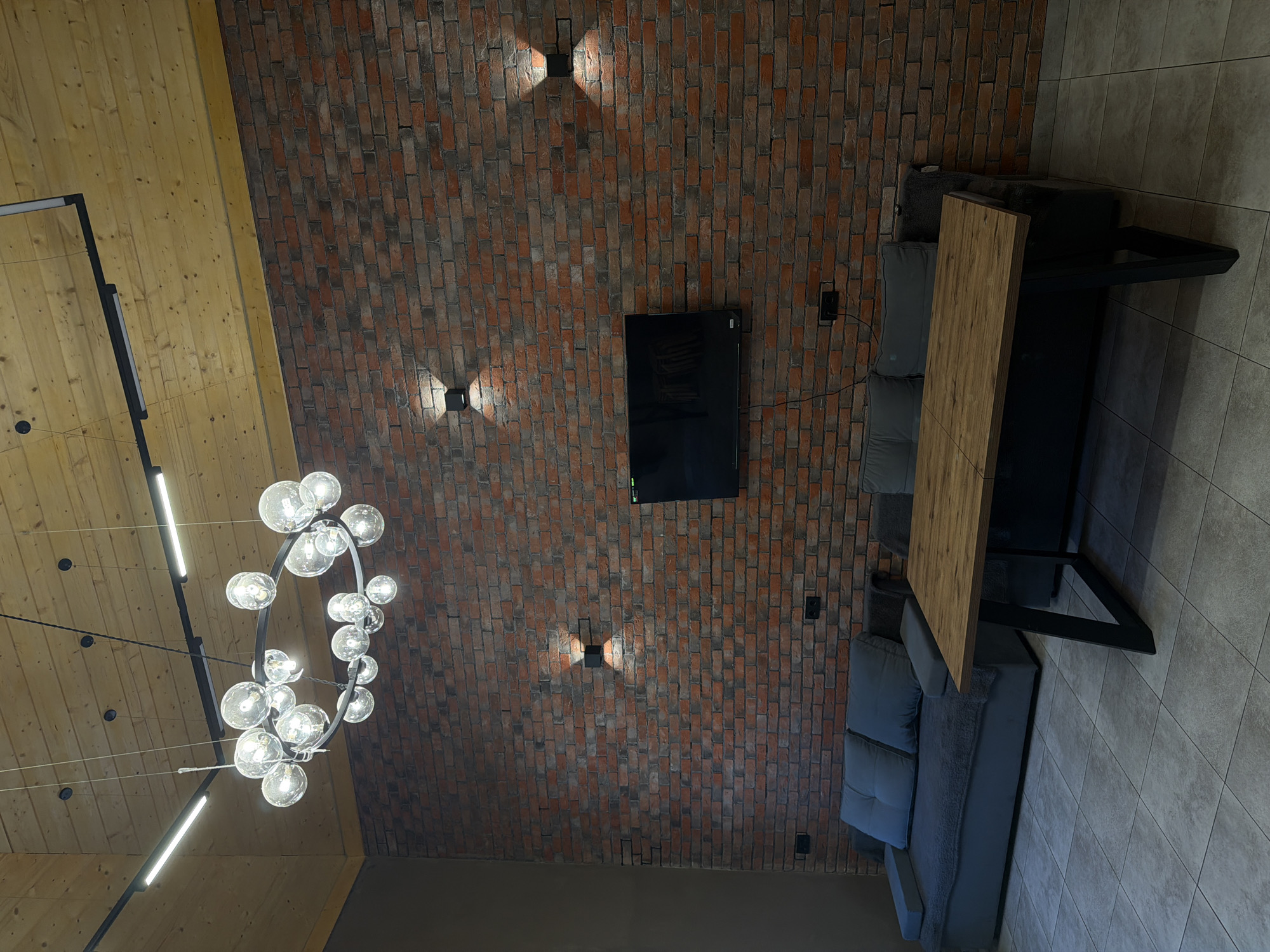 The image shows an interior space with a mix of textures and materials. The ceiling is divided between wooden planks and a brick wall. A modern chandelier hangs from the wooden ceiling. The brick wall features a flat-screen TV and four square sconce lights. A grey sectional couch and a table with a wooden top and black metal legs are positioned near the brick wall. The floor is covered in grey tiles. The location is possibly Kamennoye, Russia, based on the prompt. No people are visible.