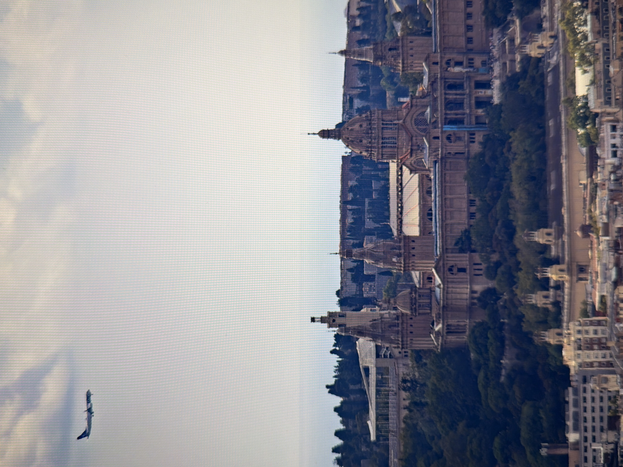 The image shows a cityscape with a large, ornate building in the foreground, appearing to be a palace or museum. The building has multiple domes, towers, and classical architectural elements. It is situated on a hill covered with trees. In the background, there is a clear sky with a few clouds and an airplane flying in the distance. A portion of a modern city with smaller buildings is visible at the bottom of the image. The scene appears to be a panoramic view of a historical district within a larger urban environment. The location is not Kyiv, Ukraine, but rather likely Barcelona, Spain, given the architecture which resembles the Palau Nacional on Montjuïc.