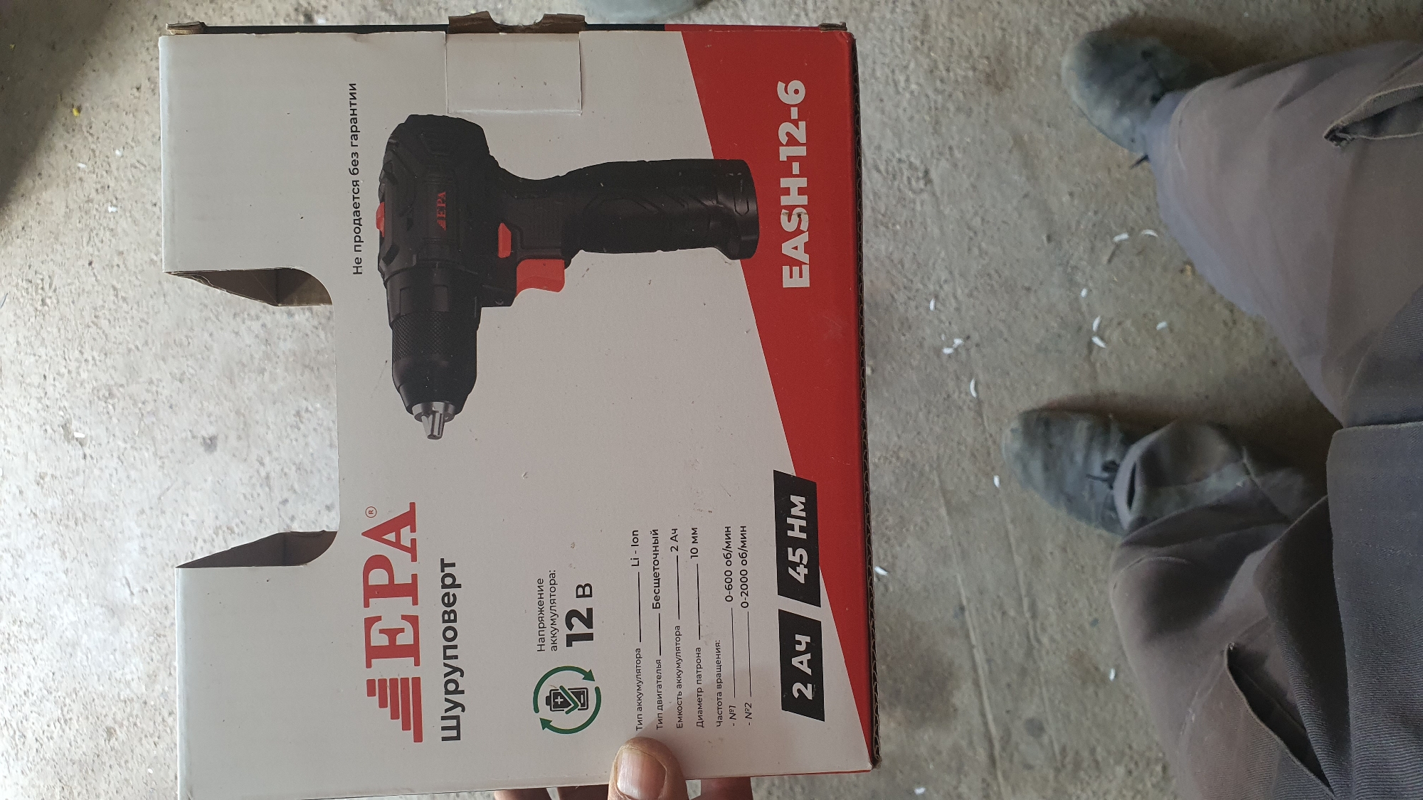 An overhead, medium shot captures a cardboard box containing an EPA brand electric screwdriver, positioned on a concrete floor. The box, predominantly white with red accents, displays product specifications including "12V" for voltage, "Li-ion" for battery type, and "45 Nm" for torque. The screwdriver itself is black with red and gray accents, featuring the EPA logo. In the foreground, the lower legs and feet of an individual wearing brown pants and dark, worn boots are visible, suggesting their presence and likely interaction with the product. The surrounding environment is a rough, speckled concrete floor with scattered white debris.