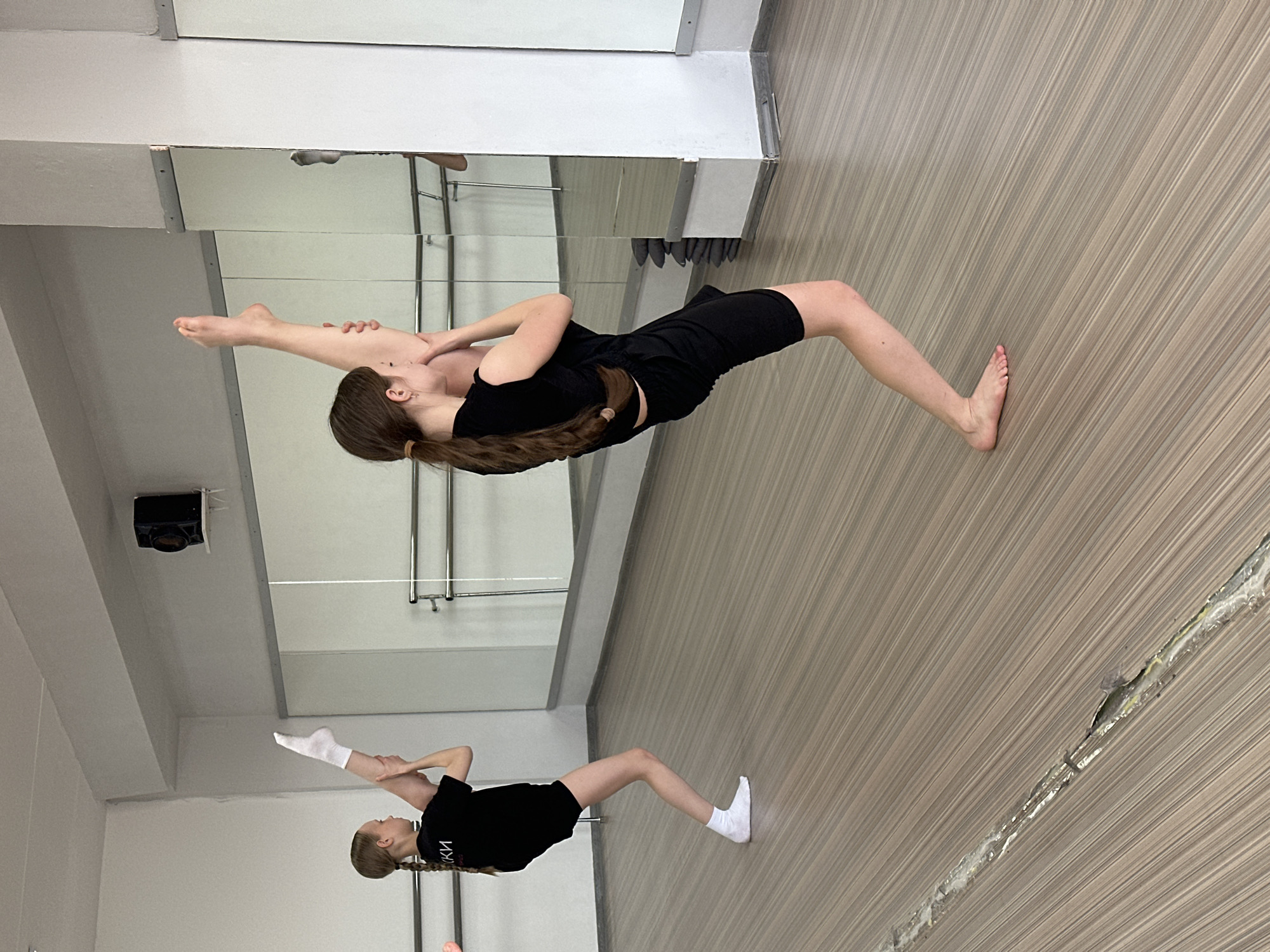 The image shows a young woman in a dance studio. She is wearing a black t-shirt and black shorts. Her hair is in a ponytail. She is holding one leg straight up in the air. She is in front of a mirrored wall. The reflection shows a similar pose with white socks on the feet. The floor is wooden. The studio is located in Votkinsk, Russia.