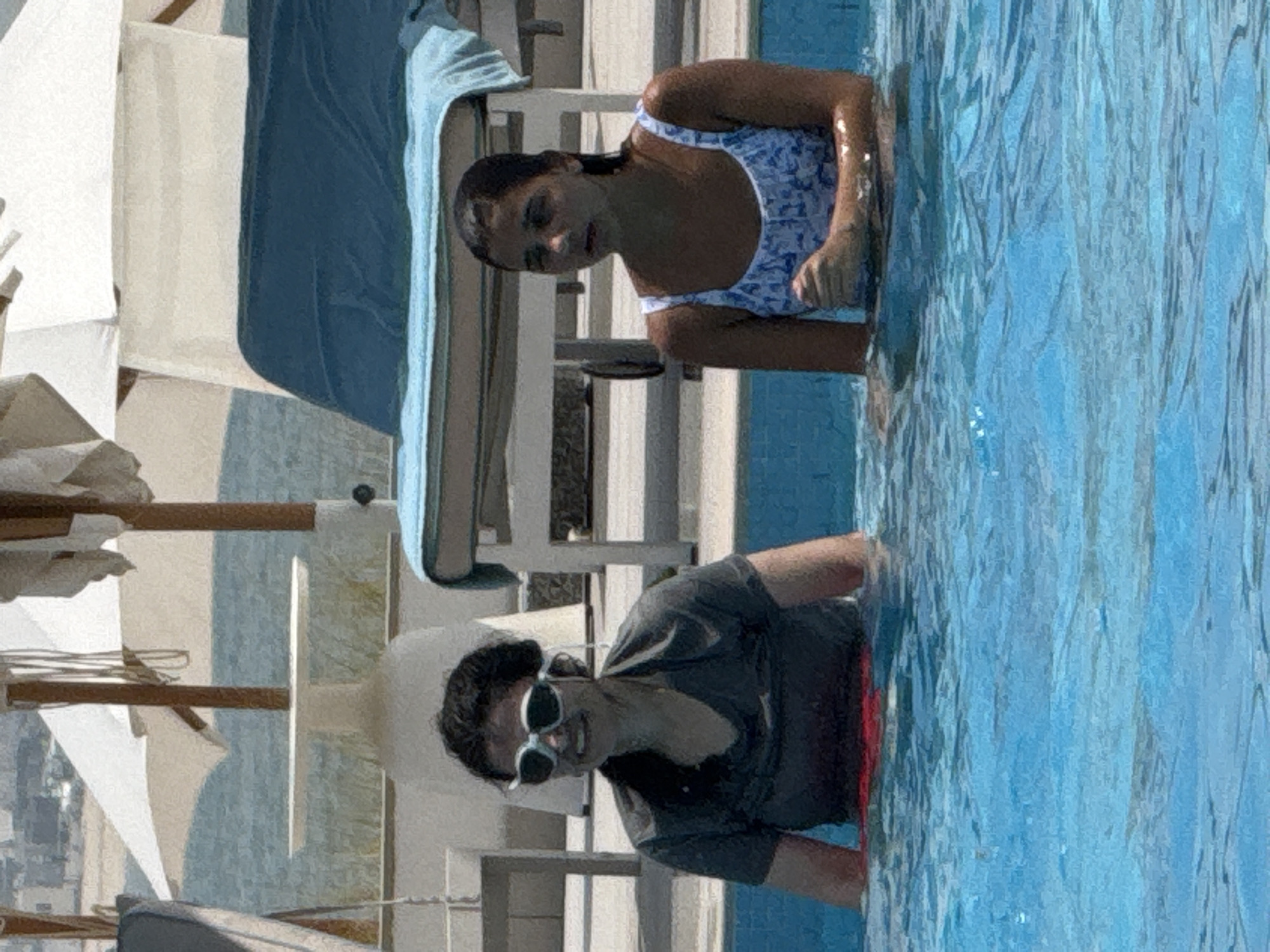Two females are in a swimming pool. The female on the left, wearing a dark grey t-shirt and sunglasses, is looking towards the camera and smiling. The female on the right, wearing a white and blue patterned swimsuit, is looking forward with a neutral expression. The pool is filled with blue water, and the edges of the pool are visible. In the background, there are lounge chairs, umbrellas, and a body of water, suggesting a resort or hotel setting in a city.