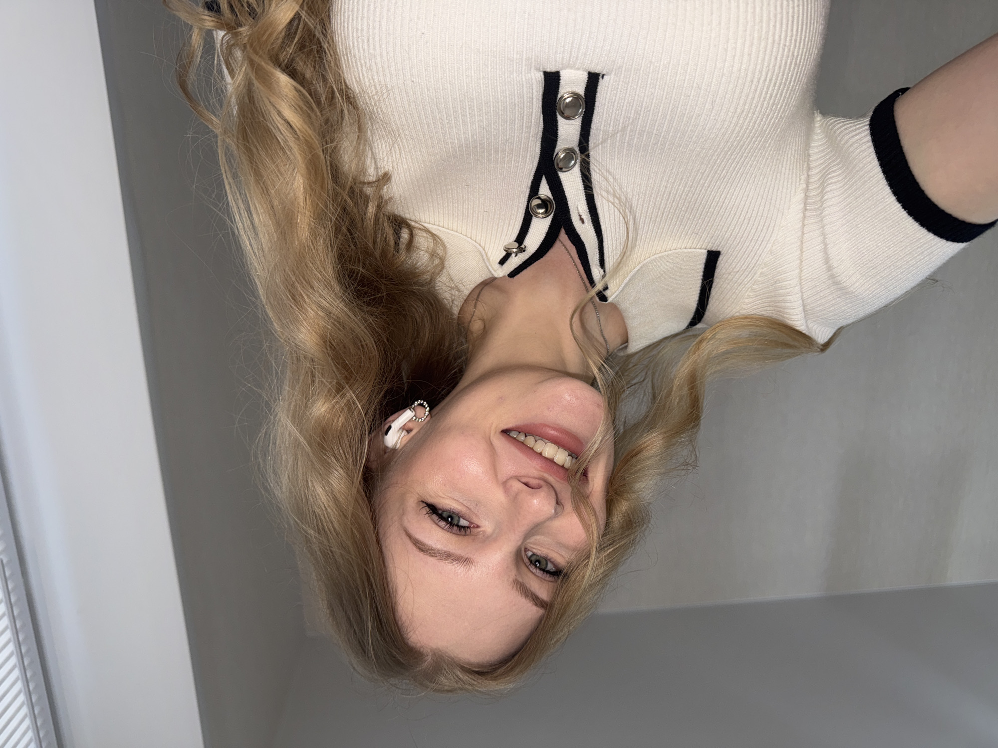 A fair-skinned woman with light blonde hair poses for a photo. Her hair is styled in loose waves, with some strands falling across her face. She is smiling, revealing her teeth. She wears a white ribbed top with black trim and silver buttons. In her left ear, she has a white wireless earbud. The background consists of a light gray wall, and a window can be seen on the left side. The location is unknown, and there is no indication of the city of Perm, Russia.