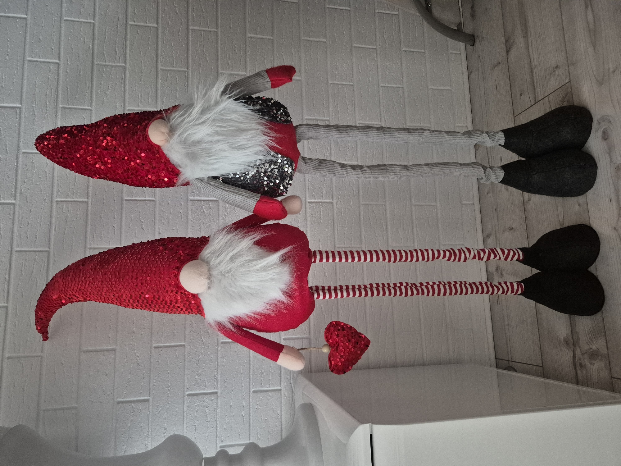 The image shows two decorative gnomes standing in front of a white brick wall. The gnome on the left is wearing a red sequined hat, a red tunic, and striped red and white leggings. It is holding a red sequined heart in its left hand. The gnome on the right is wearing a red sequined hat, a gray vest with silver sequins, and gray ribbed leggings. Both gnomes have white beards and pink noses. They are standing on a wooden floor. The background consists of a white brick wall and a white cabinet on the left side of the image. There is no indication that this scene is located in Barbing, Germany.
