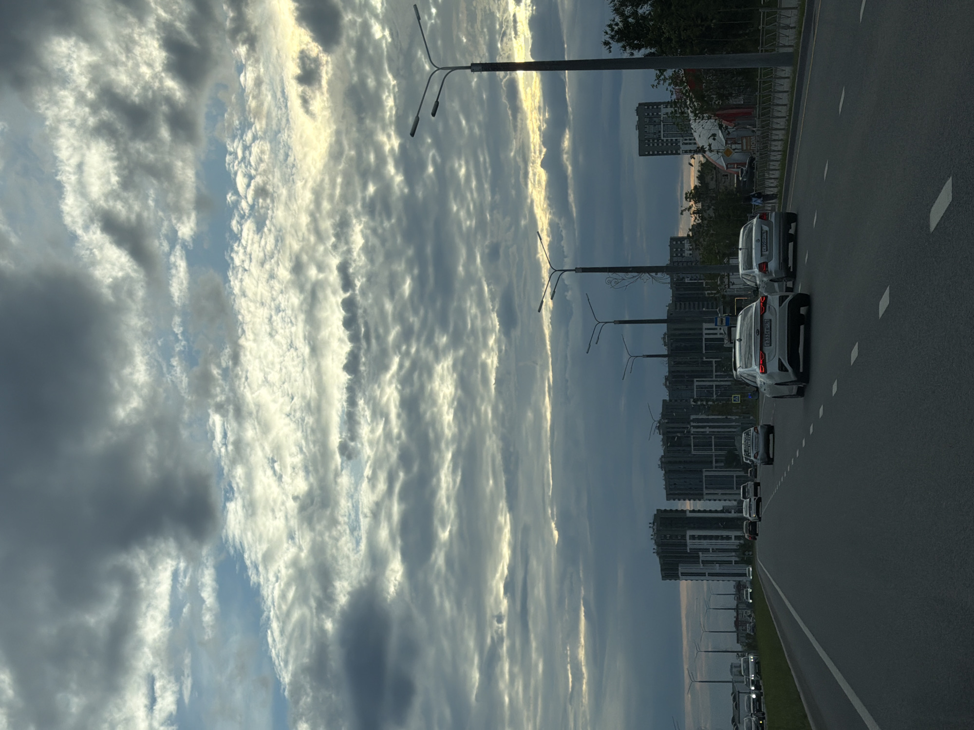 The image depicts a street scene in Kazan, Russia, with several cars driving on a multi-lane asphalt road. The perspective is from a vehicle traveling along the road, looking forward. The sky is filled with dramatic, textured clouds, some dark and ominous, others illuminated by sunlight, suggesting it might be late afternoon or early evening. Tall modern buildings, characteristic of urban development, line the right side of the road in the distance. Streetlights are visible along the roadside. The overall impression is one of movement and urban life under a dynamic sky. There are no discernible people in the image, only vehicles. The license plates on the cars are not clearly legible for specific details.