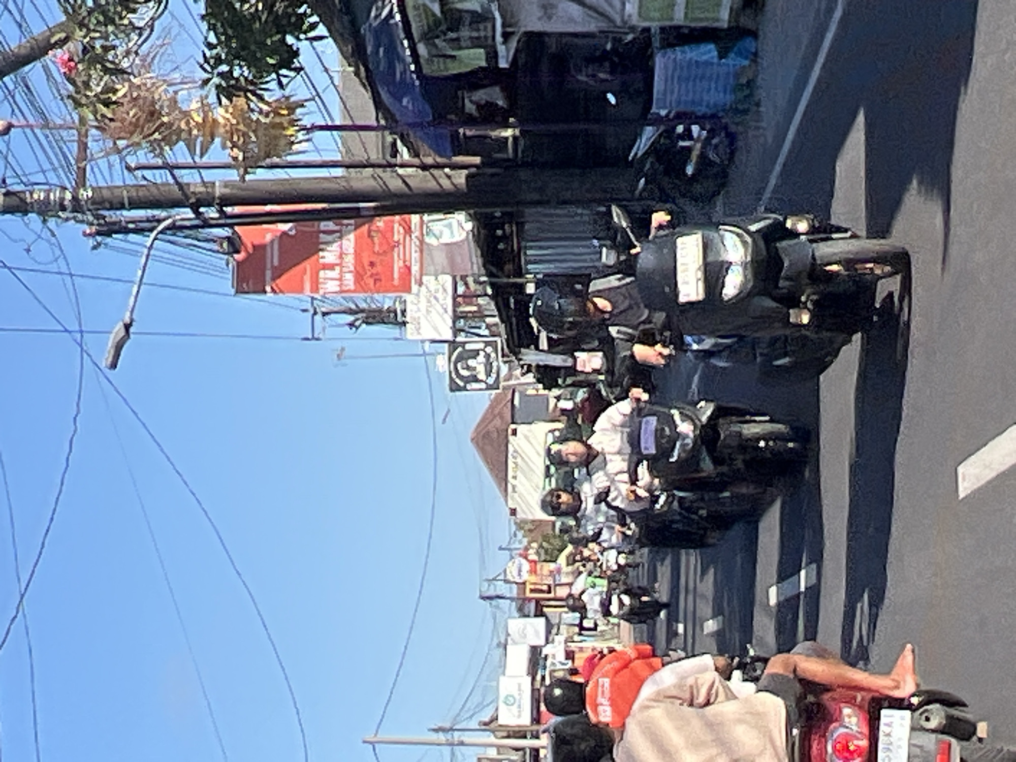 The image shows a street scene in Denpasar, Indonesia, with a procession of motorcycles on a paved road. The motorcycles are traveling in the same direction, with several riders wearing helmets. Some riders are wearing casual clothing, while others are in brighter, possibly uniform attire. The road is marked with white lines. On the right side of the road, there are buildings with awnings, some appearing to be small shops or stalls. A prominent red billboard with white text is visible in the background, and a utility pole with numerous wires is also present. Trees and foliage can be seen, suggesting an outdoor urban or semi-urban environment. The sky is clear and blue, indicating daytime. The overall atmosphere is that of a busy street with motorized traffic.