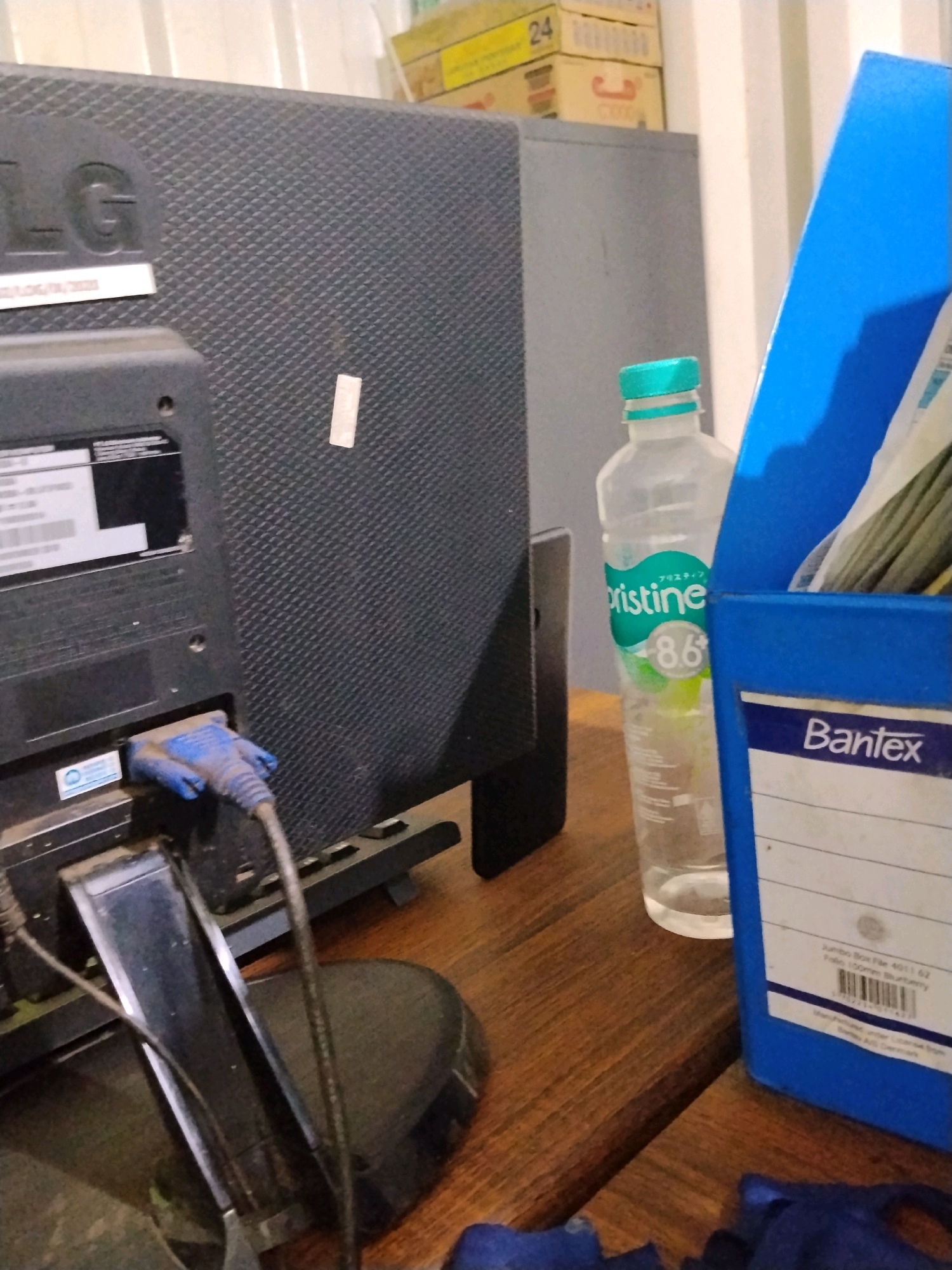 This is a forensic, matter-of-fact description of a scene in Sempayau, Indonesia. The image displays a desk with a computer monitor, its stand, and various cables. A blue binder labeled "Bantex" is positioned on the right side of the desk. Next to the binder is a partially filled plastic bottle of "Pristine" water. In the background, stacked boxes are visible, with one box displaying the number "24". The overall environment appears to be an indoor setting with artificial lighting.