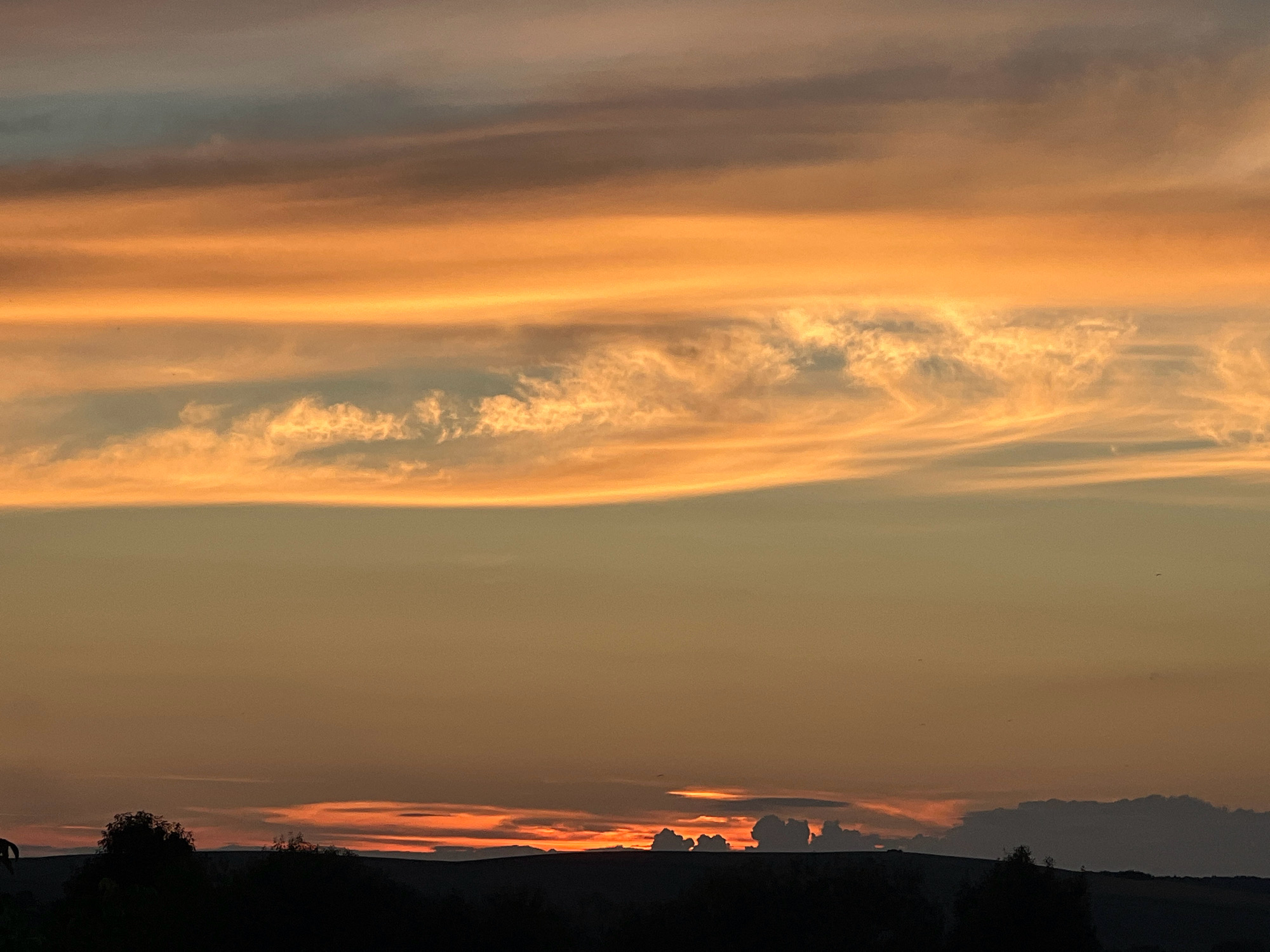 The image depicts a sunset over Horodyshche, Ukraine. The sky is filled with dramatic clouds painted in vibrant hues of orange, yellow, and muted gray. The sun, partially obscured by the horizon, casts a warm glow across the sky. In the foreground, a silhouetted landscape of rolling hills and sparse trees creates a dark contrast against the luminous sky. There are no people or discernible actions visible in the image. The scene is peaceful and evocative of the natural beauty of the Ukrainian countryside at dusk.