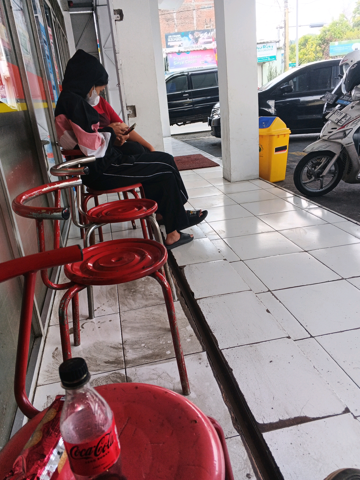 A medium-shot photo shows a person in a black hoodie and black pants sitting on a red chair, looking at their phone. The person is wearing a white face mask. To their left, another person is partially visible, also sitting on a red chair. In the background, there are two black cars parked and a white motorcycle. A yellow trash can is visible next to a white pillar. The ground is tiled with white and gray squares. A partially consumed bottle of Coca-Cola Zero Sugar is on a red chair in the foreground. The scene appears to be an outdoor waiting area in Genuk Barat, Indonesia, possibly at a business or service center.