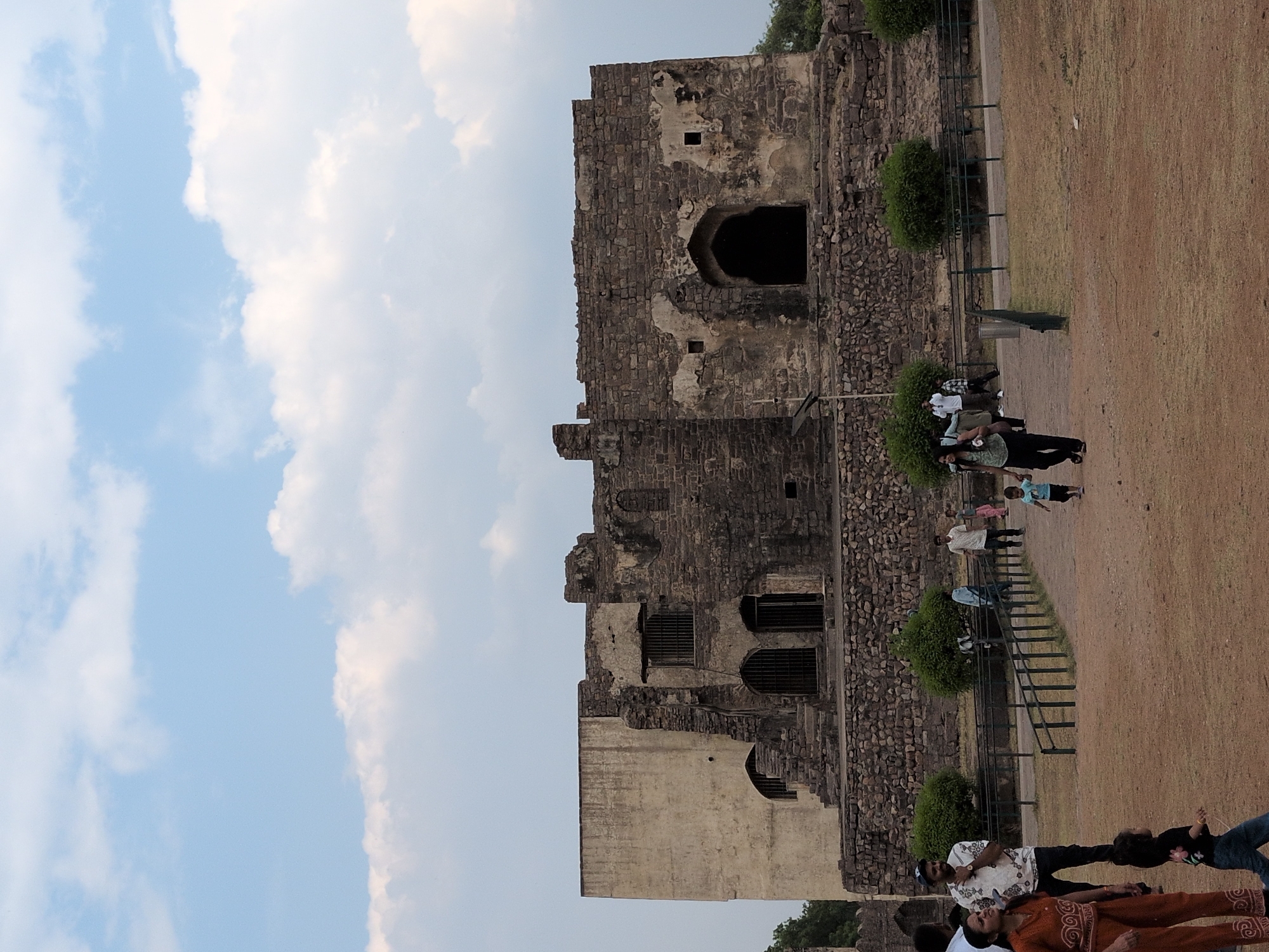 This image depicts a section of the Golconda Fort in Hyderabad, India, under a light blue sky with scattered white clouds. The primary subject is a large, multi-level stone structure exhibiting characteristics of ancient architecture and significant wear. The left portion of the structure has a smoother, lighter-colored, possibly stuccoed wall with a visible arched opening, while the right and central portions consist of dark, rough-hewn stone blocks, indicating varying stages of construction or degradation. This darker section features multiple arched doorways and barred windows, with a large, dark, central arched entrance on the lower level. Patches of crumbling material are visible on the stone walls.
In the foreground, a reddish-brown dirt path occupies the lower half of the image. A dark metal fence runs horizontally across the mid-ground, separating the path from the area directly in front of the stone structure. Several rounded green bushes are visible behind the fence line. Approximately ten individuals, including adults and children, are present in the lower half of the scene. On the left foreground, a woman in an orange-brown outfit stands facing left, with a small child in a black shirt and blue jeans walking to her right. In the center, a man in a dark shirt carries a small child on his shoulders while walking right, accompanied by another small child in a blue shirt walking ahead of them. Further back, near the fence, other adults and children are walking or standing, including a man in a patterned white shirt and another individual wearing a light blue head covering.