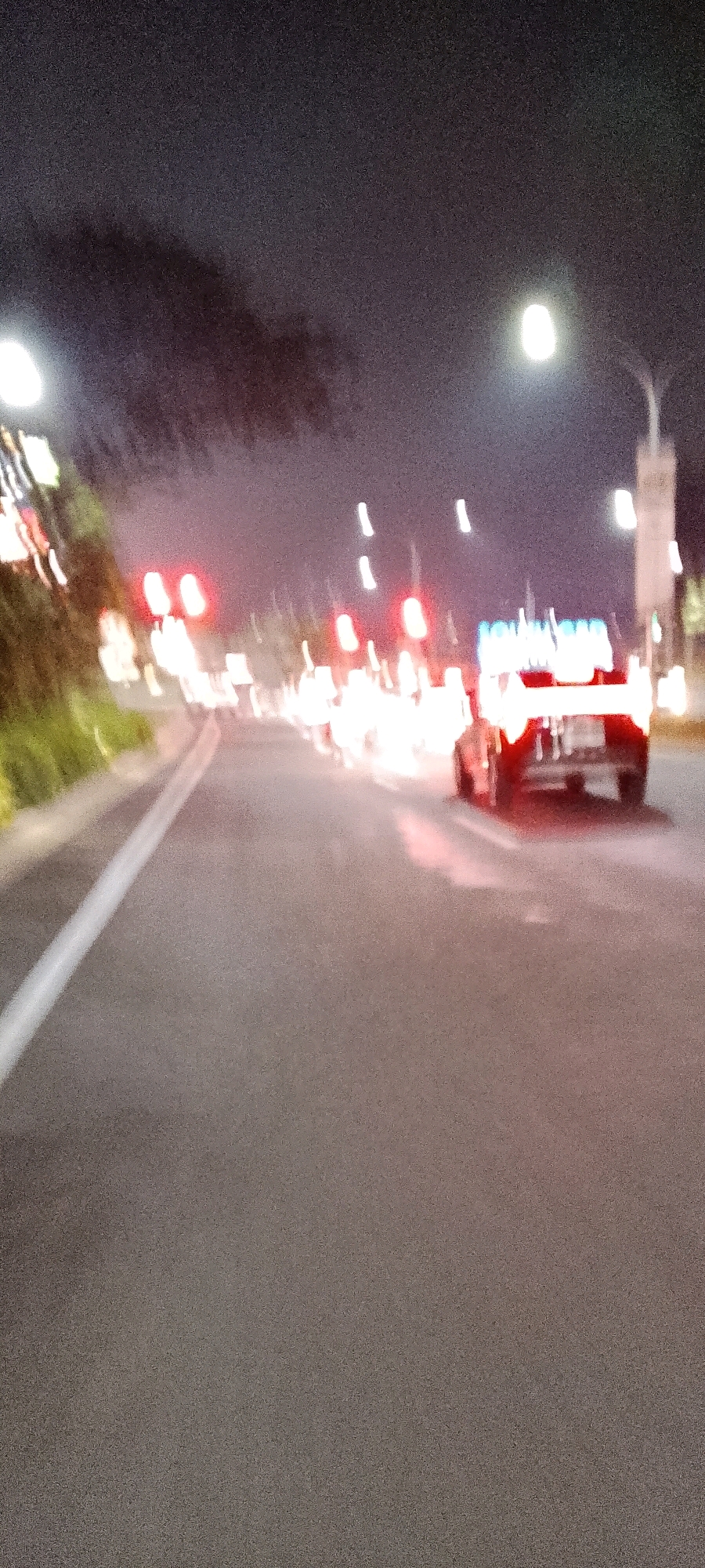 The image depicts a blurry, nighttime scene on a paved road, likely in Gunung Putri, Indonesia. A dark-colored car is visible in the right lane, its taillights illuminated red. Other red and white lights, possibly from traffic signals or other vehicles, are scattered throughout the scene. Trees with dark foliage are present on the left side of the road, with some green vegetation visible at their base. A white line marks the edge of the road in the foreground. The sky is dark and appears to have some grainy texture. The overall impression is of a street with moderate traffic and urban lighting at night.