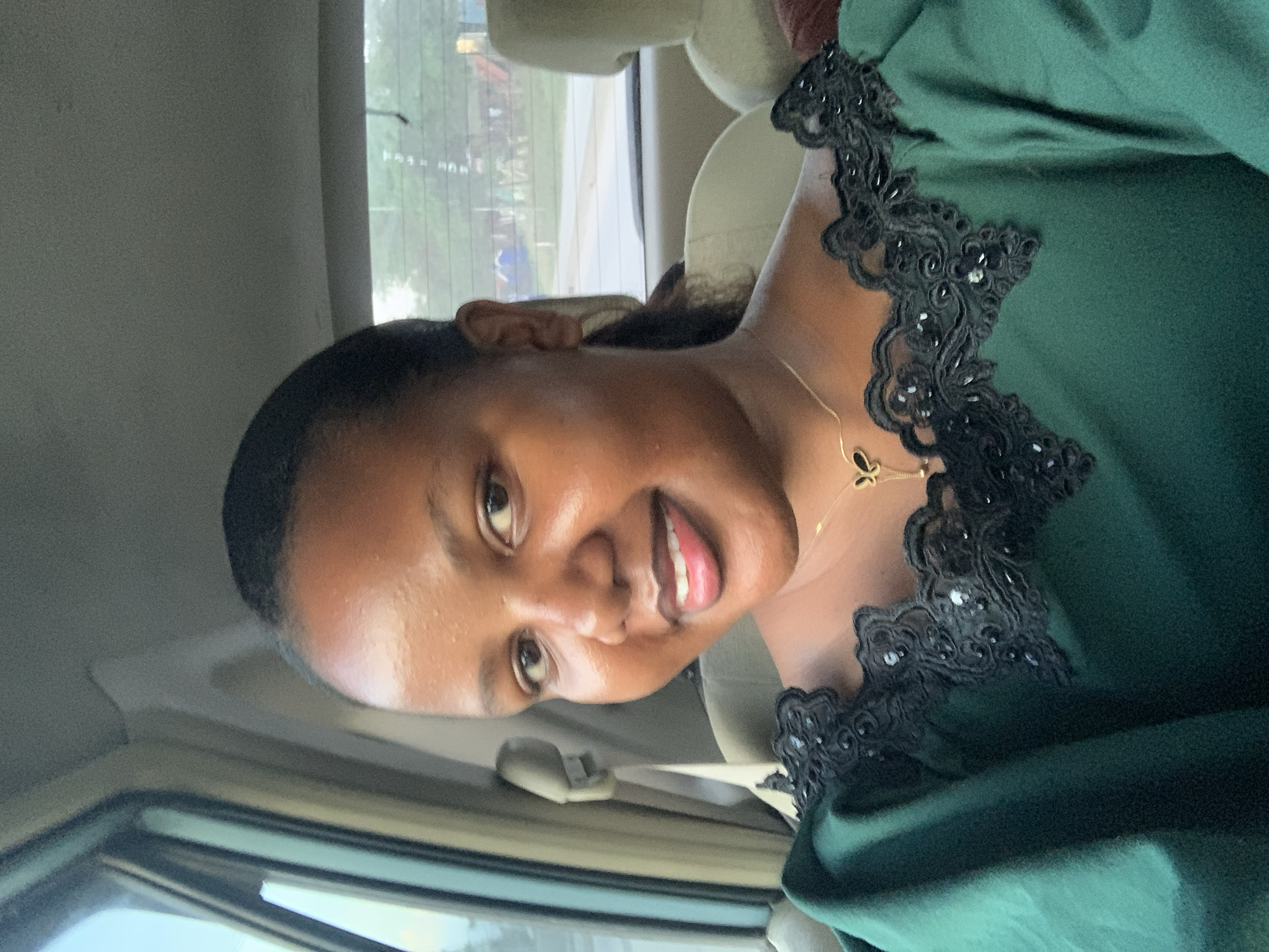A medium, angled shot shows a woman inside a vehicle, possibly a car. The woman has dark skin and short, dark hair, and she is wearing a green top or dress with black lace detailing around the neckline. She is also wearing a gold necklace. The interior of the vehicle is visible, including the seat belt and the window. Outside the window, a blurred view of a city street or road can be seen, likely in Dar es-Salaam, Tanzania.