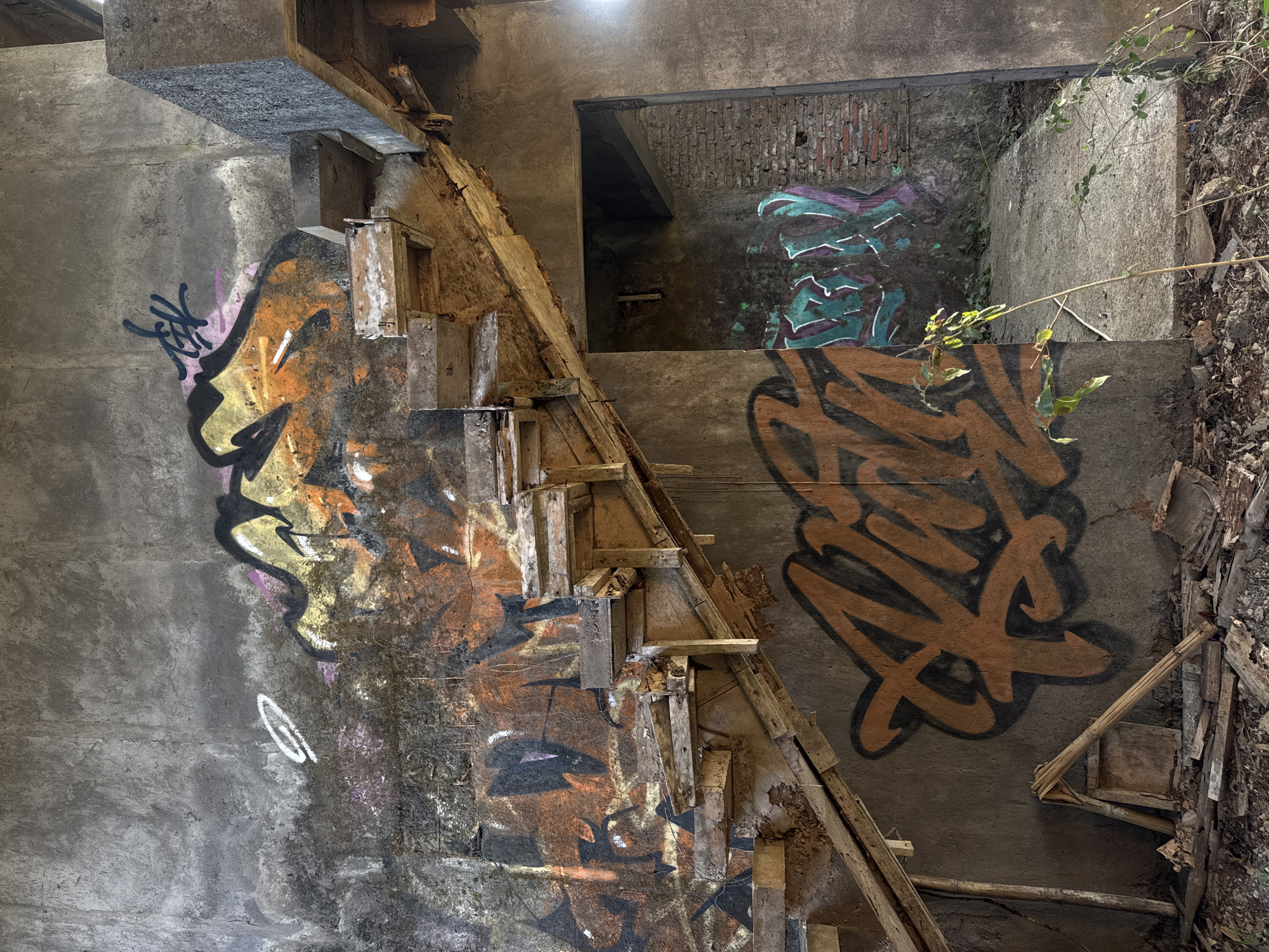 The media file shows a dilapidated concrete structure, likely an abandoned building, with graffiti art adorning its walls. The scene depicts a derelict staircase made of wood and concrete, partially collapsed and covered in debris. The walls are covered in vibrant graffiti tags and artistic pieces, predominantly in shades of orange, black, purple, and green. A large tag in orange with a black outline is prominently displayed on the right side of the image. On the left side, a graffiti piece appears to depict a stylized face or figure in orange, yellow, and black.
The setting suggests an outdoor or semi-exposed environment, with plants and dry leaves visible on the ground and creeping up the structure, indicating a period of neglect and the passage of time. The lighting is natural, suggesting daylight, but the overall atmosphere is one of decay and abandonment. There is no visible text beyond the graffiti. The location context of Kuta Selatan, Indonesia, implies a tropical or subtropical environment, which aligns with the presence of plants and the weathered appearance of the structure. No people are present in the image.