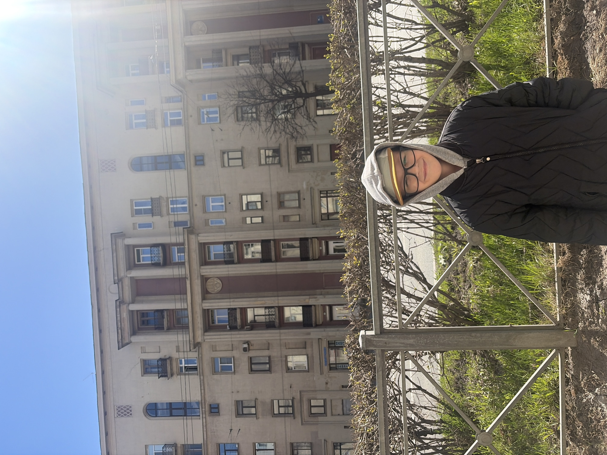 A person wearing a black quilted jacket and a grey hooded sweatshirt stands outside a building in Saint Petersburg, Russia. The building behind them is multi-storied, with rows of rectangular and arched windows, decorative moldings, and a pale facade. A metal fence runs parallel to the person, with grass and small trees visible behind it. The sky is clear blue.