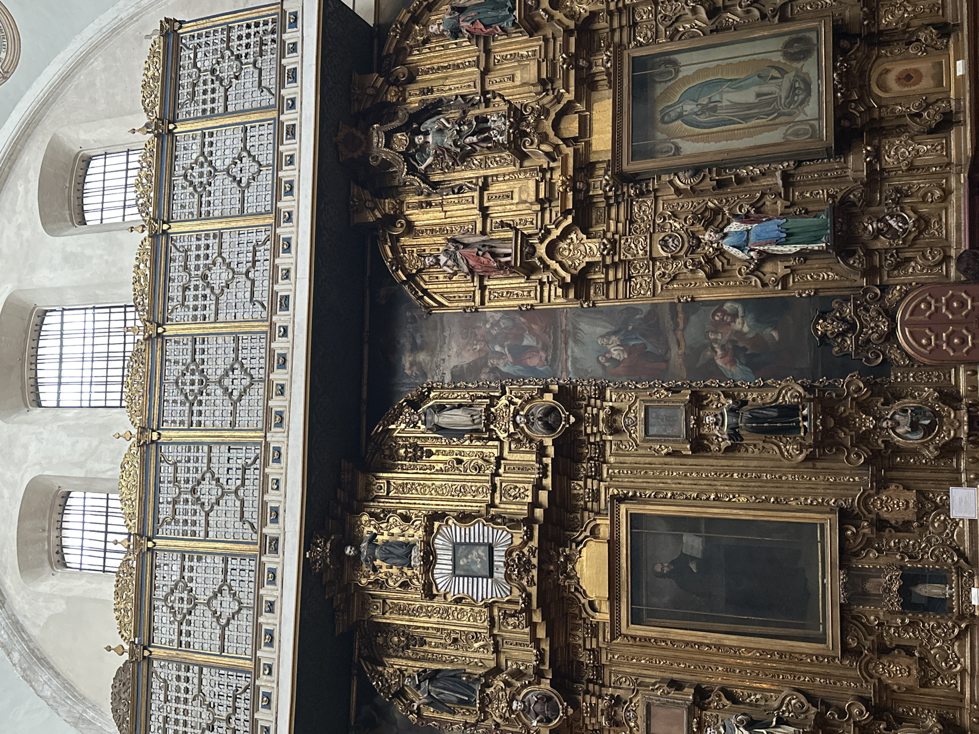 This image depicts an ornate interior, likely a church, in Mexico City, Mexico. The photograph is oriented at a slight angle. The left side shows a tall, white wall with three arched windows that have barred grilles. Attached to this wall is a highly decorated, geometric structure with gold trim and blue detailing. To the right is a wall adorned with intricate gold carvings and several framed paintings or images. A larger mural or painting stretches across the center of the gold wall. The architecture and decoration are indicative of Baroque or Colonial styles. There are some human figures depicted in the mural and statues on the carved gold wall.