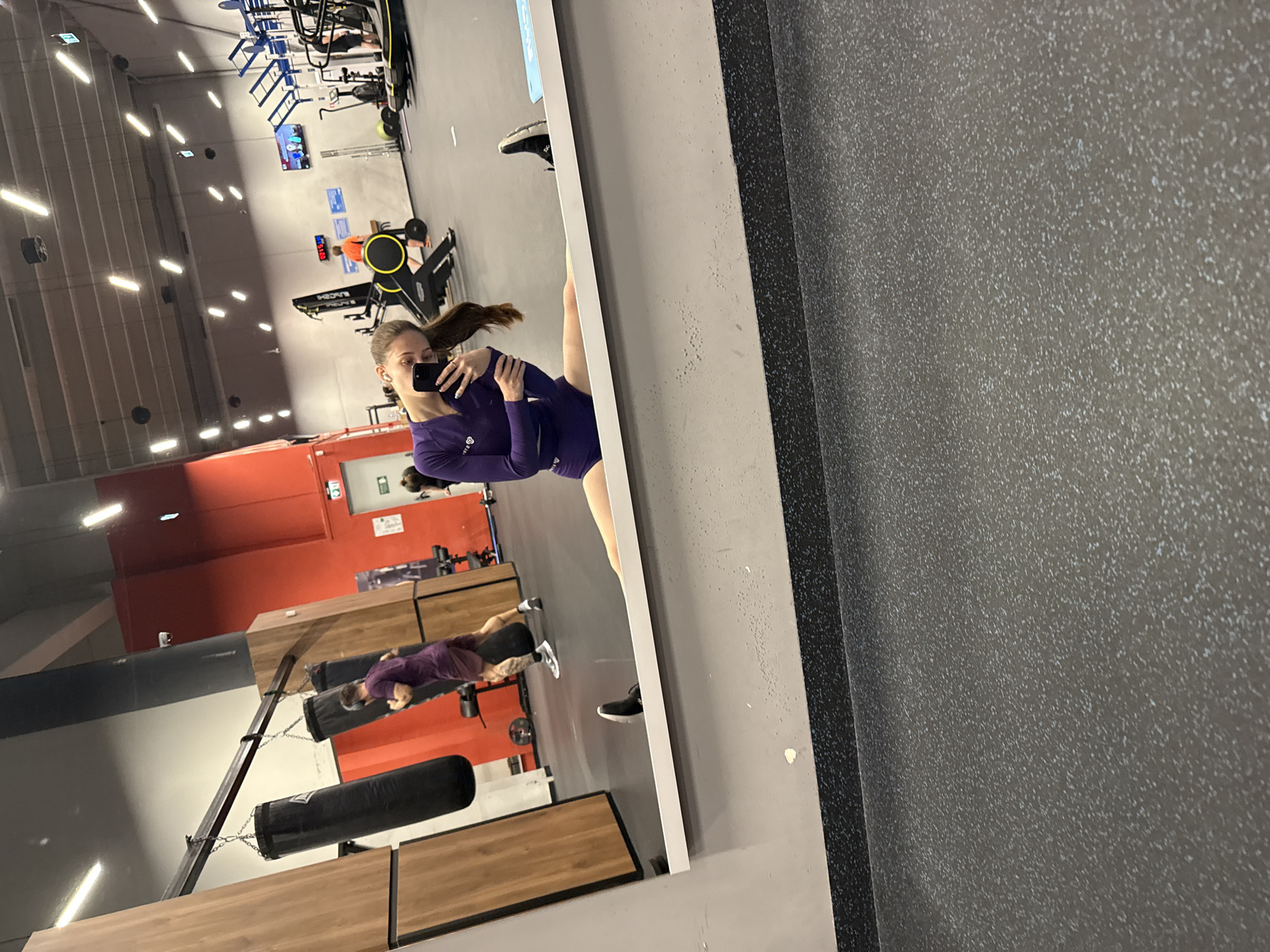 A person is holding a phone to take a picture in a mirror inside a gym. The person is wearing a long-sleeve purple shirt and matching bottoms. In the reflection, other people are working out on various gym equipment. There are punching bags in the background and a red wall. The location is likely a gym in Sofia, Bulgaria.