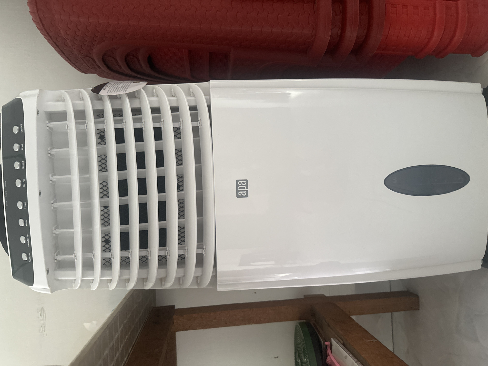 The image shows a white evaporative cooler, or air cooler, positioned indoors next to a wooden table on the left and a stack of red plastic containers on the right. The cooler has a black control panel on top with various buttons and indicators, including labels for "Ionizer," "Humidifier," "Mode," "Timer," "Swing," "Speed," "Cool," and "ON/OFF." The main body of the cooler is white, with a slatted front section for air dispersion and a dark, oval-shaped water level indicator window towards the bottom. A tag with text is attached to the side of the cooler. The floor appears to be tiled. The presence of a wooden table, tiled flooring, and stacked plastic containers suggests a domestic or commercial storage area. The image does not provide specific visual cues to pinpoint its location to the city of Binjai, Indonesia.