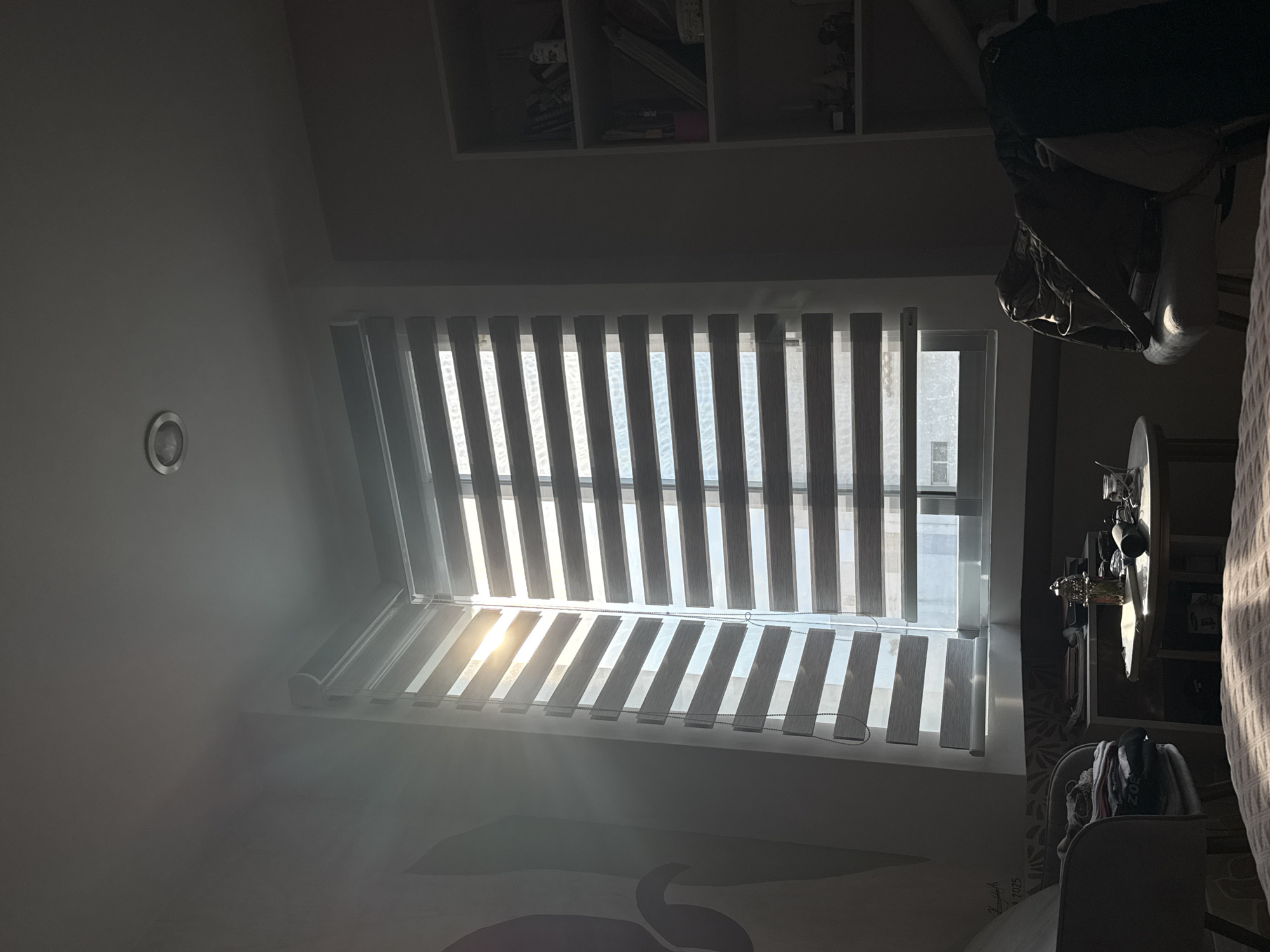 An interior scene in San Bernardino Tlaxcalancingo, Mexico, featuring a window with open vertical blinds allowing sunlight to stream into the room. Sunlight is visible coming through a window with blinds. A shelf with assorted items sits near the top of the frame, and items hang from a rack nearby. The room is somewhat dark, with strong sunlight.