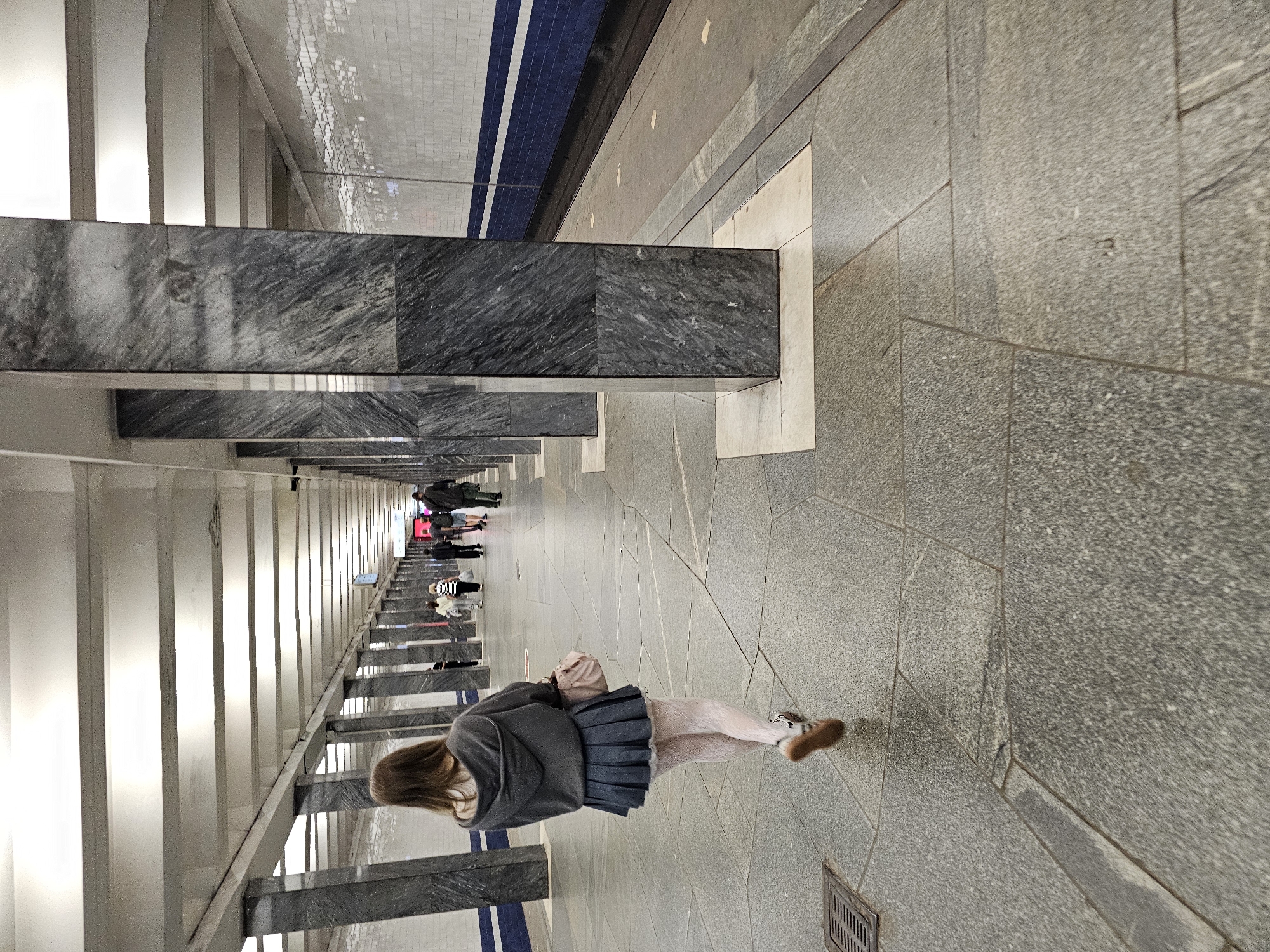 The image depicts a pedestrian in a subway station in Moscow, Russia. A woman, seen from behind, walks down a platform. She is wearing a grey hooded jacket, a dark pleated skirt, and white patterned stockings. She carries a light pink handbag. The subway platform is characterized by large, dark marble pillars, a tiled wall with blue and white stripes, and a granite-tiled floor. Other pedestrians are visible in the background, further down the platform. The lighting is bright, suggesting an indoor environment.