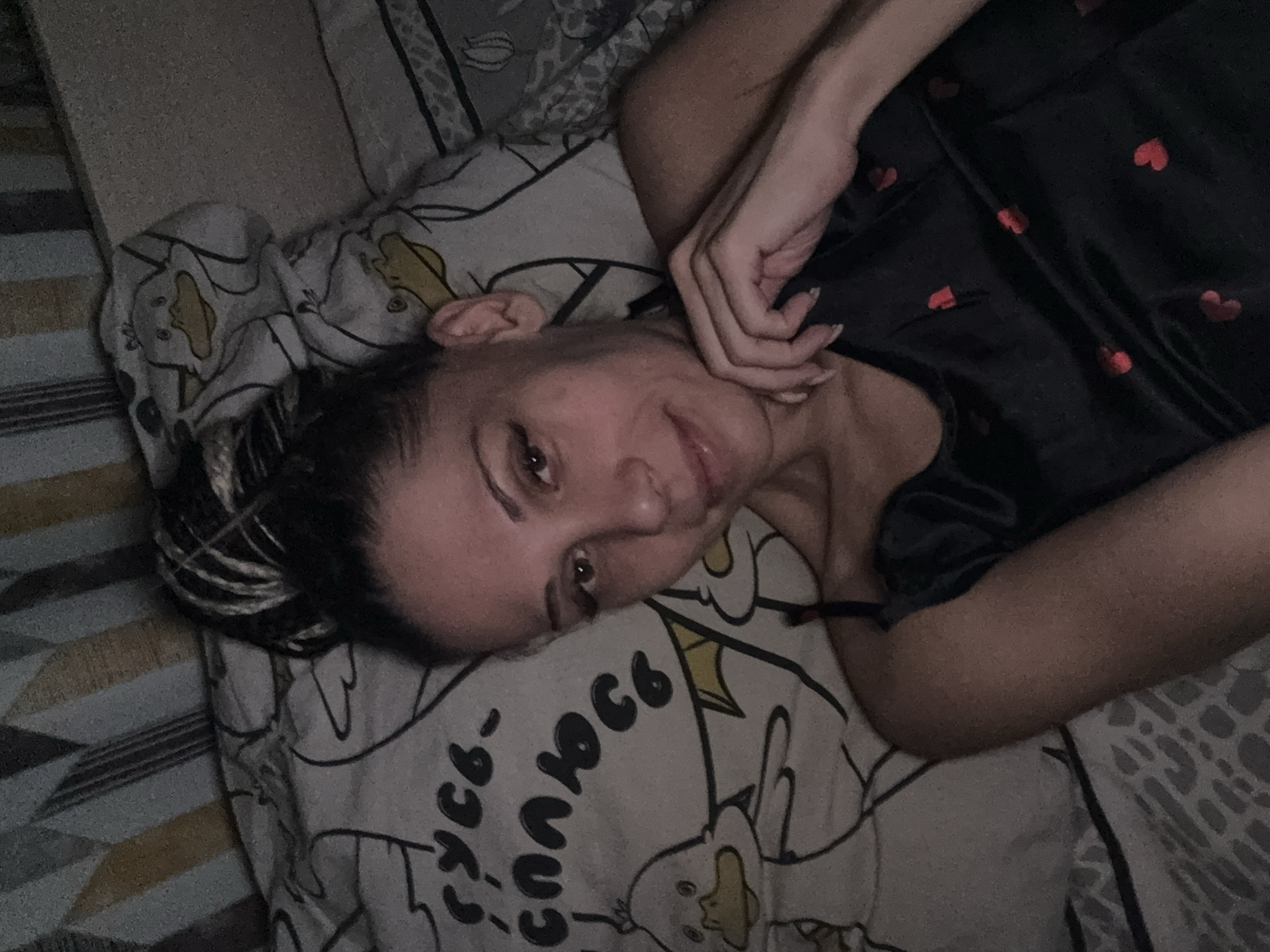 A woman is lying on a bed. She has her head tilted on a patterned pillow. The woman's hand is near her face. She is wearing a dark-colored top with a heart pattern. The location is unknown.