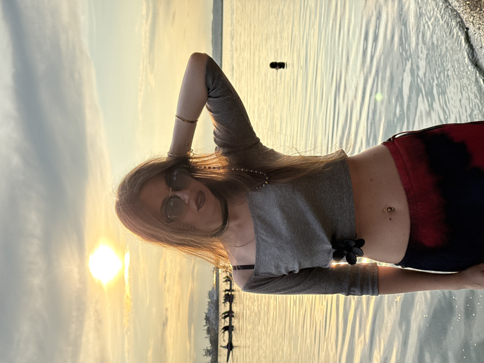 A young woman stands on the edge of a body of water, facing the camera at an eye-level shot. The sun is setting behind her, casting a warm, golden glow across the scene and reflecting off the water. She has long, wavy brown hair and is wearing sunglasses, a grey off-the-shoulder cropped top with a black flower accent, and dark pants with a red tie-dye pattern. Her right arm is bent and resting on the back of her head, with her left hand also near her head. In the background, there are several figures in boats on the water, and a distant shoreline with industrial structures. The overall atmosphere is serene and picturesque.