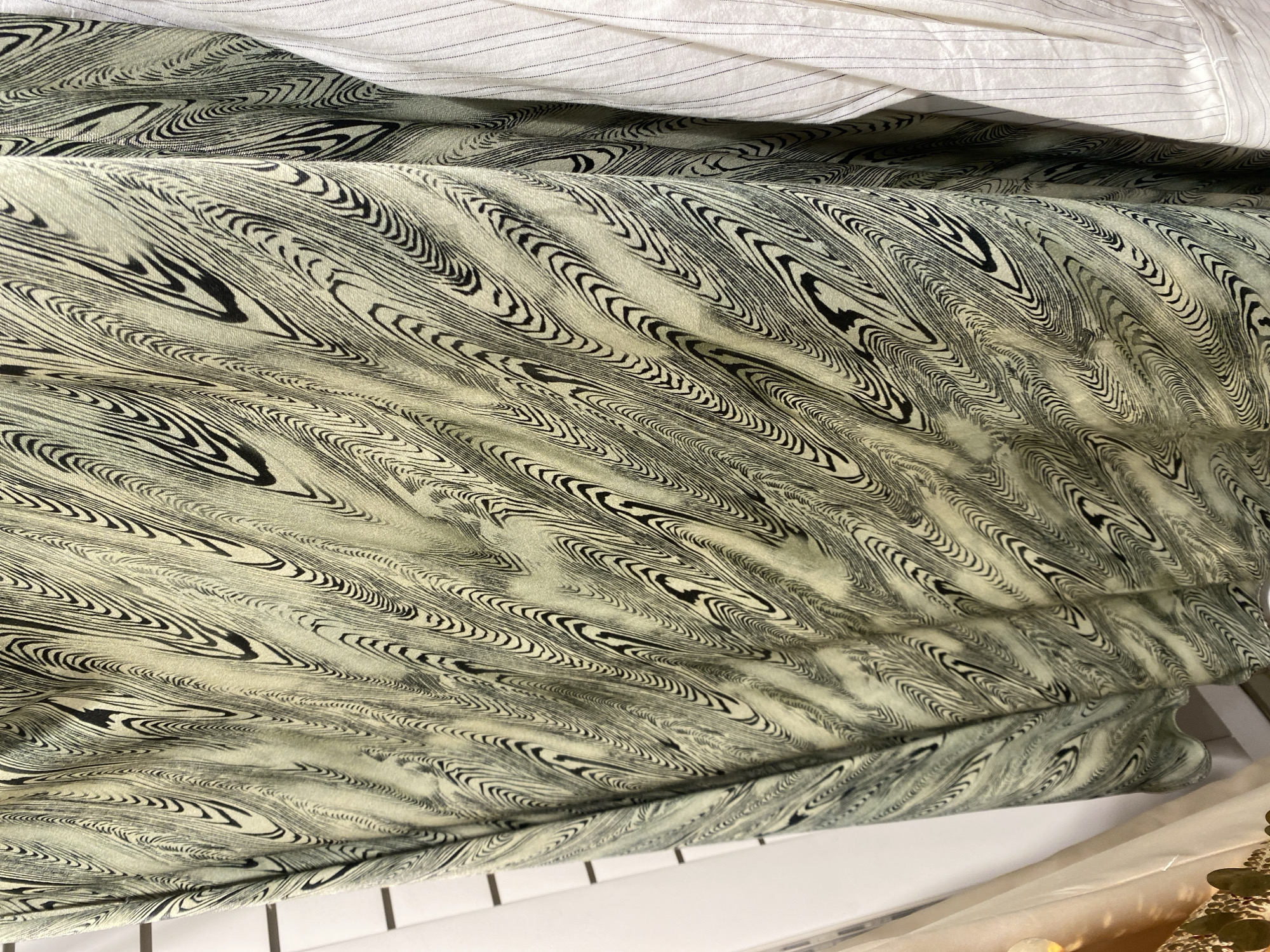 The image primarily displays textiles, with a central focus on a fabric featuring a distinctive abstract pattern. This main fabric has a light, muted background color, appearing as a pale greenish-beige or off-white, adorned with intricate, flowing lines in black. The pattern consists of numerous swirling, wave-like, and irregular concentric shapes, reminiscent of a marbled effect or dense wood grain. The fabric is draped and folded, creating soft undulations and shadows that enhance its texture and the fluid quality of the design.
In the upper section of the frame, a portion of a second fabric is visible. This textile is white with very fine, subtle black pinstripes running vertically, and it appears to be folded or gathered. Below the patterned fabric, a flat, white surface is present, exhibiting thin black vertical lines that suggest a tiled or paneled structure, possibly a countertop or shelf. In the bottom right corner, a decorative element can be partially seen, appearing to be a cluster of green, possibly artificial, leaves interspersed with reflective or glittery accents. The overall lighting is soft and diffused, casting gentle shadows and suggesting an indoor setting. No human subjects, specific activities, or visible text are present. The atmosphere is calm and focused on the textures and patterns of the materials.