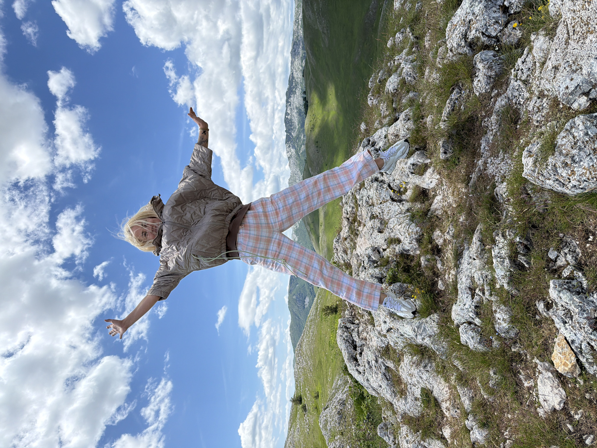 A full-body shot captures a young individual, likely female, with blonde hair, mid-jump on a rocky outcrop in Novakovići, Montenegro. The person is centrally positioned, with arms outstretched to the sides, hands open, and hair appearing to fly upwards due to the motion. They are wearing a light brown or tan puffy short-sleeved jacket and light-colored plaid pants featuring shades of pink, white, and light blue. White athletic shoes are visible on their feet.
The immediate foreground consists of a rugged, light-colored rocky terrain interspersed with patches of green grass and occasional small yellow wildflowers. Beyond the foreground, the midground and background reveal expansive rolling green hills under a bright blue sky dotted with numerous large, fluffy white clouds. Distant mountains with varying textures and shades of green and grey are visible on the horizon. The scene is illuminated by bright daylight.