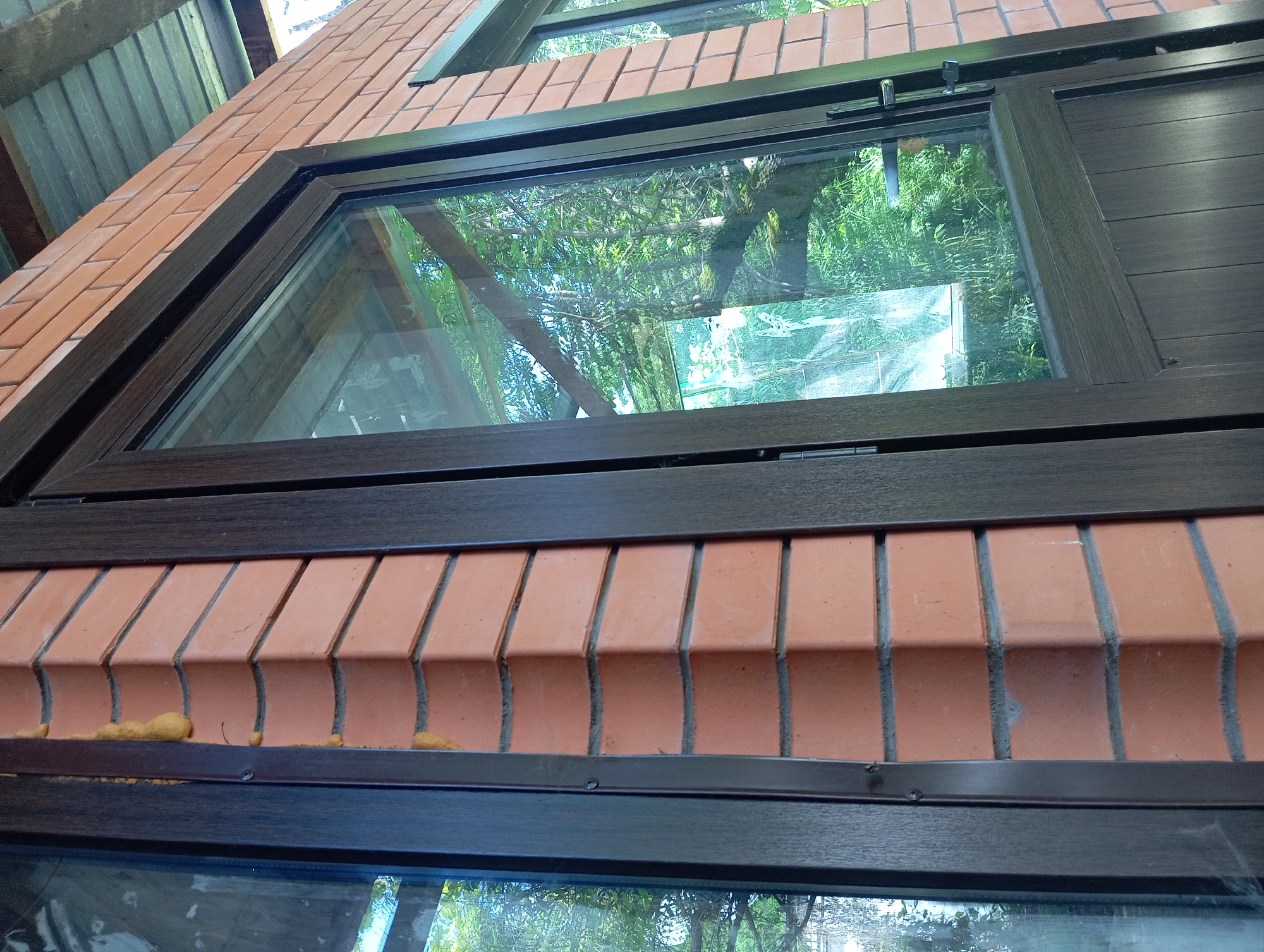 This media file shows a close-up view of a modern window installed in a brick wall. The window has a dark brown frame and the glass reflects a lush green outdoor scene with trees and foliage. The brickwork below the window is laid in a distinctive pattern, with each brick having a curved bottom edge, creating a decorative ripple effect. Small, irregular blobs of yellow material are visible along the top of this decorative brickwork, possibly insulation foam or sealant.
Above the main window, another section of the building is visible, including what appears to be part of a wooden roof structure and another window. The overall impression is of an exterior architectural detail of a building, likely a house or a modern structure, set against a backdrop of greenery. The lighting suggests it is daytime, with natural light illuminating the scene. There are no people or animals visible in the frame. The image focuses on the texture and materials of the building, specifically the brickwork and the window frame, along with the reflection in the glass. The location context is Peredovaya, Russia, but this information is not visually evident from the image itself. There is no visible text in the media file.
