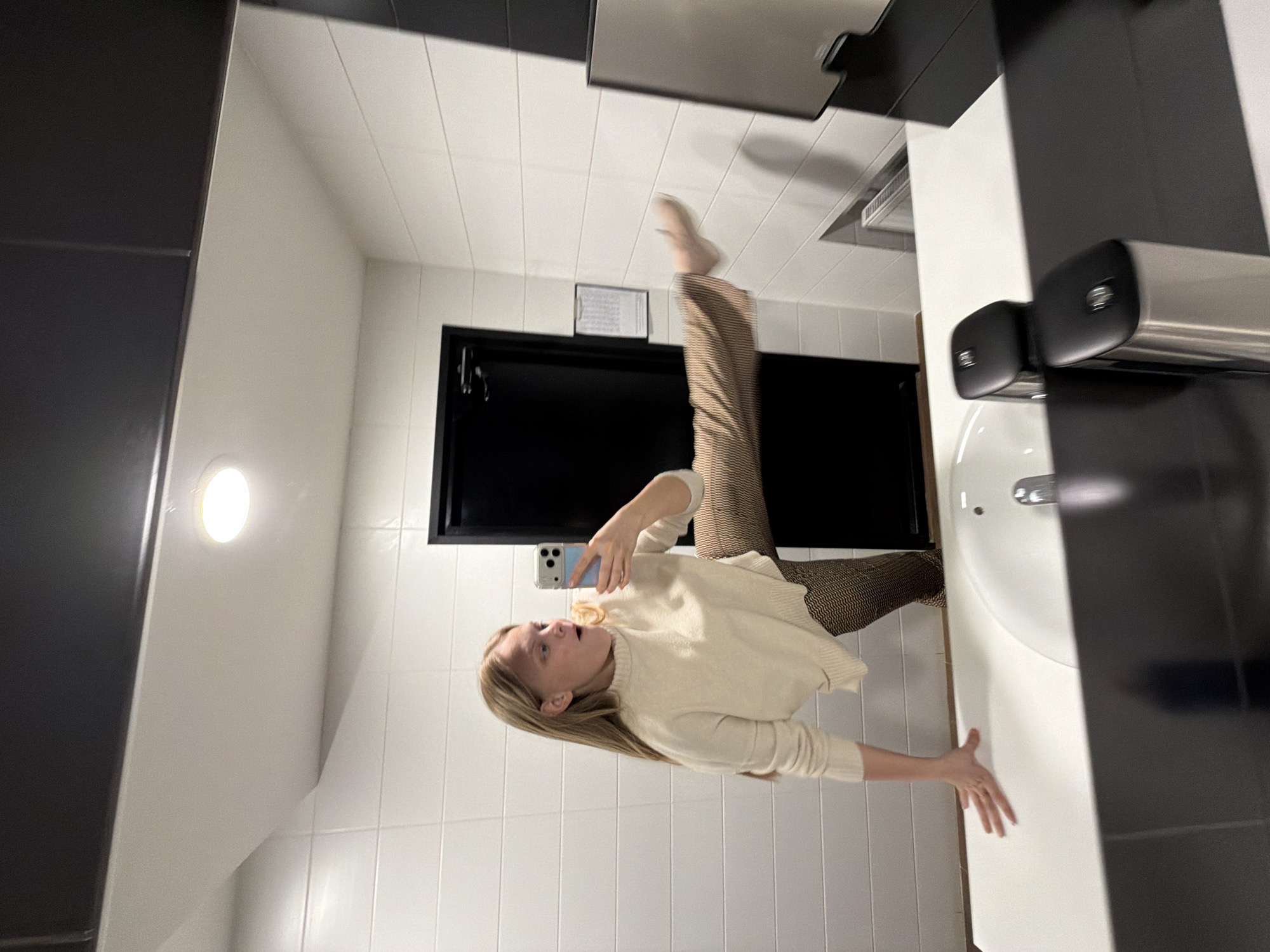 This media file captures a mirror selfie taken by a young woman in what appears to be a modern public restroom or private bathroom in Izhevsk, Russia. The image is rotated 90 degrees counter-clockwise, creating a disorienting effect, with the woman appearing to defy gravity.
The central subject is a young woman with long, straight blonde hair, wearing a light cream-colored long-sleeved sweater and brown-and-beige checkered or patterned trousers. She is seen holding a light blue smartphone in her left hand, positioned to take a photo of her reflection in the mirror. She strikes a dynamic and playful pose, balancing on one leg with her other leg lifted high into the air, bent at the knee, giving the impression she is standing on the sink counter and leaning against the wall. Her mouth is open, and her gaze is directed upwards, suggesting surprise, exertion, or an animated expression for the selfie.
The setting features clean, white tiled walls and what appears to be a white tiled ceiling. To the right of the woman is a white ceramic sink with a silver faucet. Above the sink, two modern, black and silver soap dispensers are mounted on the wall. A dark-colored door, possibly black or dark gray, is visible in the background. A circular, illuminated light fixture is affixed to the left wall. A small, rectangular piece of paper with illegible text is attached to the wall above the door. Dark gray or black panels or tiles line the upper sections of the walls. The overall impression is one of a well-lit, contemporary interior. The time of day is not discernible due to artificial lighting.